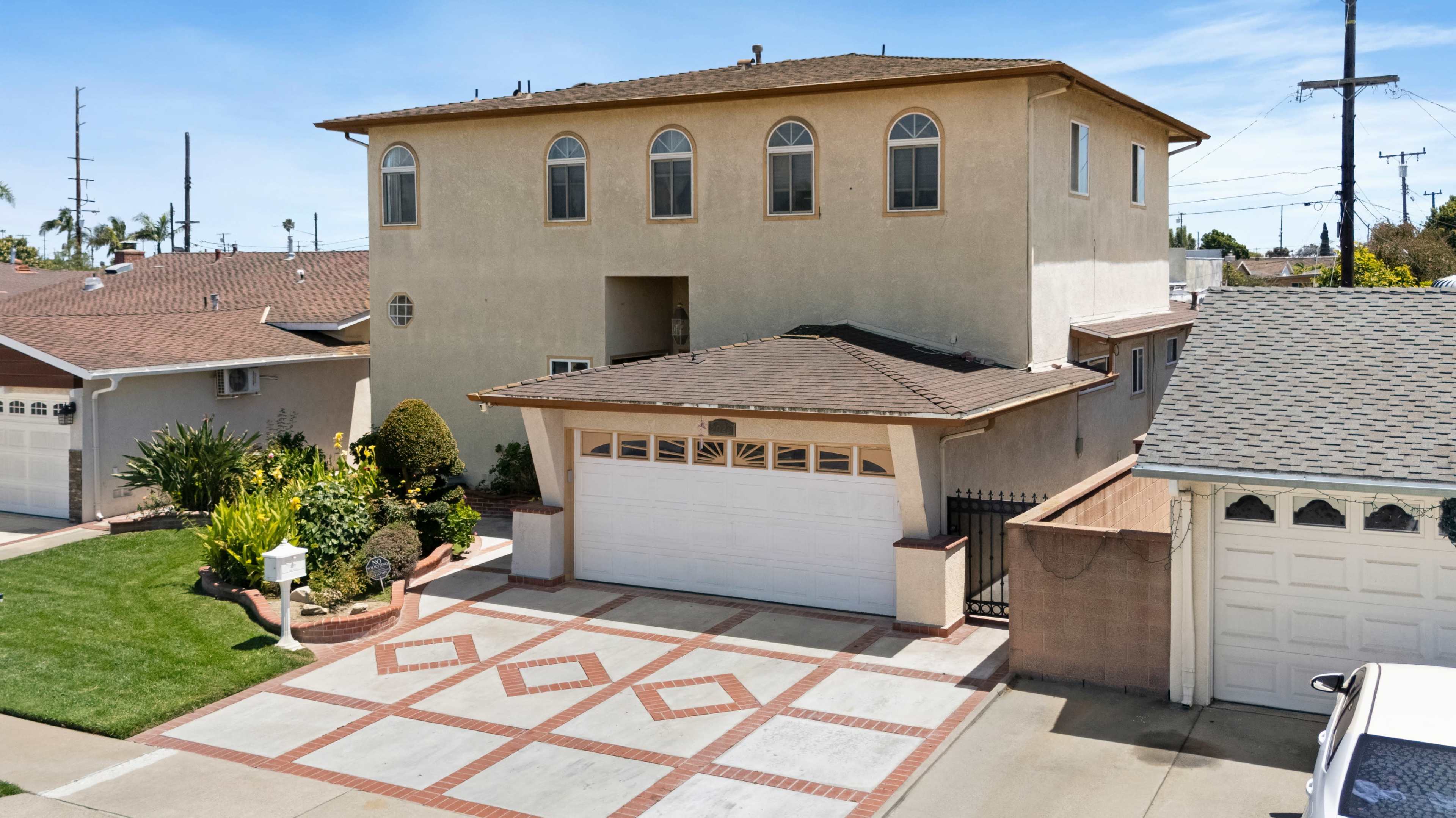 The image shows a two-story house with a garage located at the front, featuring a driveway with a patterned design and landscaped greenery nearby.