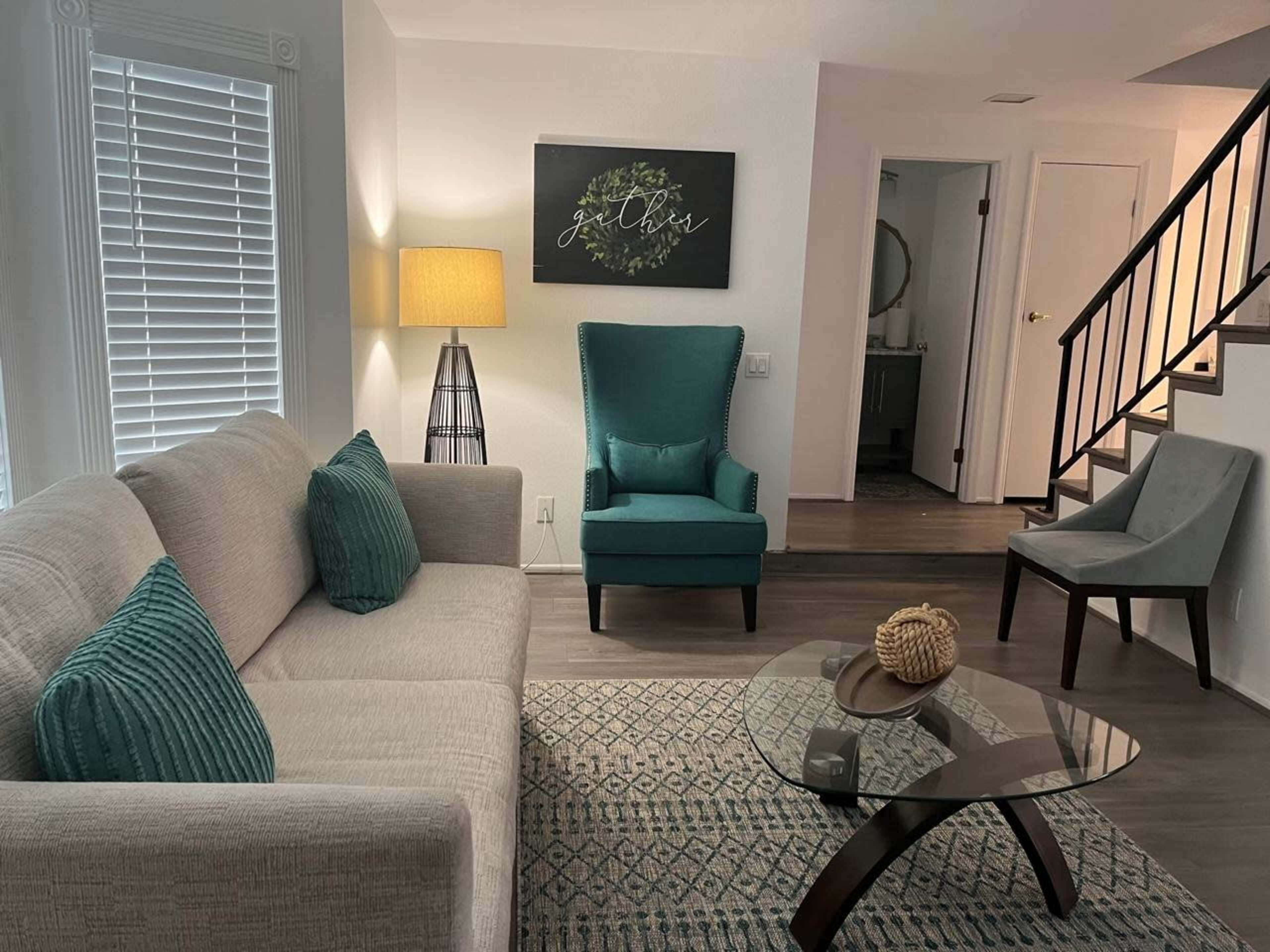 A living room features a light gray sofa with teal cushions, a glass coffee table, and modern chairs, complemented by a lamp and artwork on the wall.