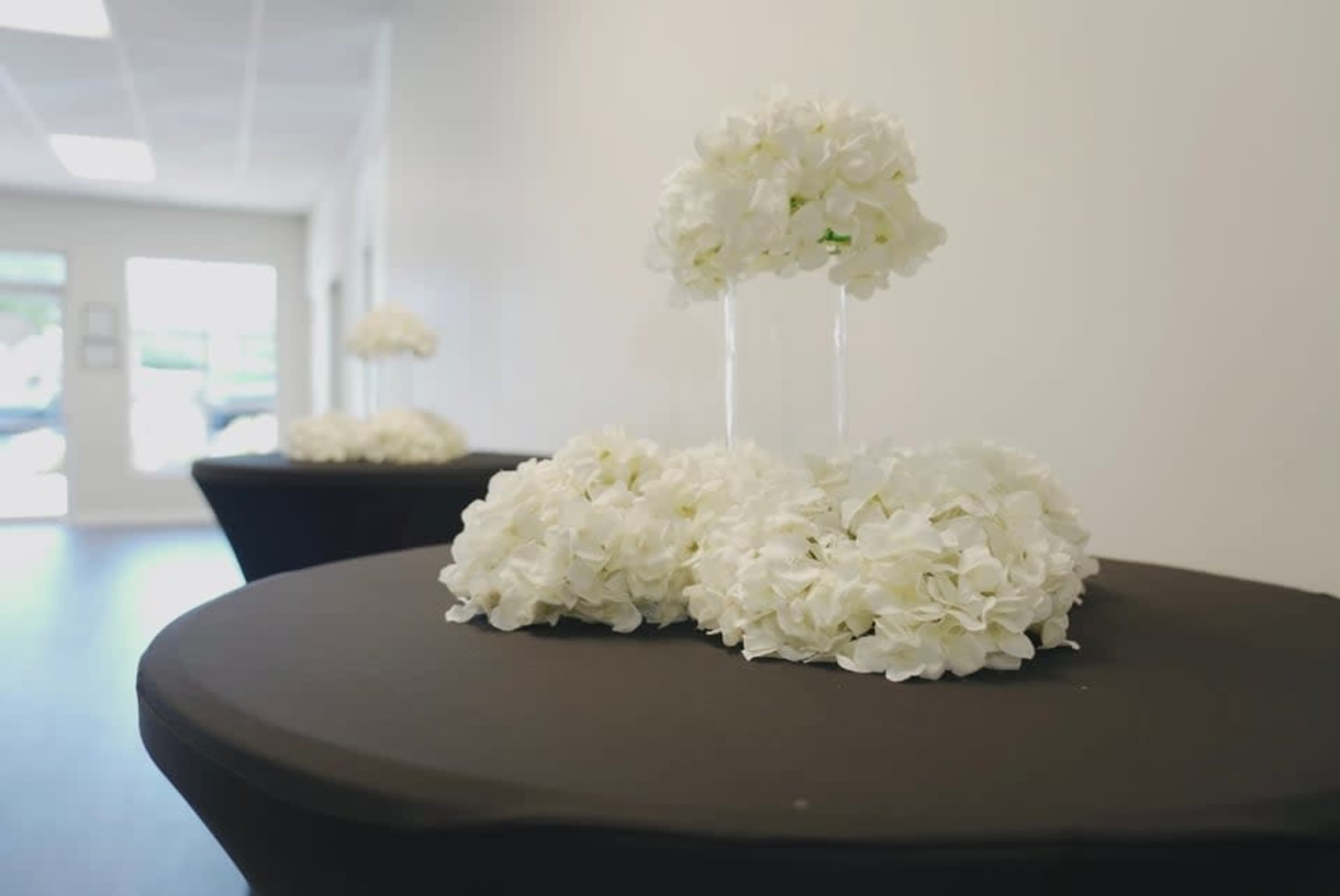 Two black tables are decorated with arrangements of white flowers, set against a plain white wall.