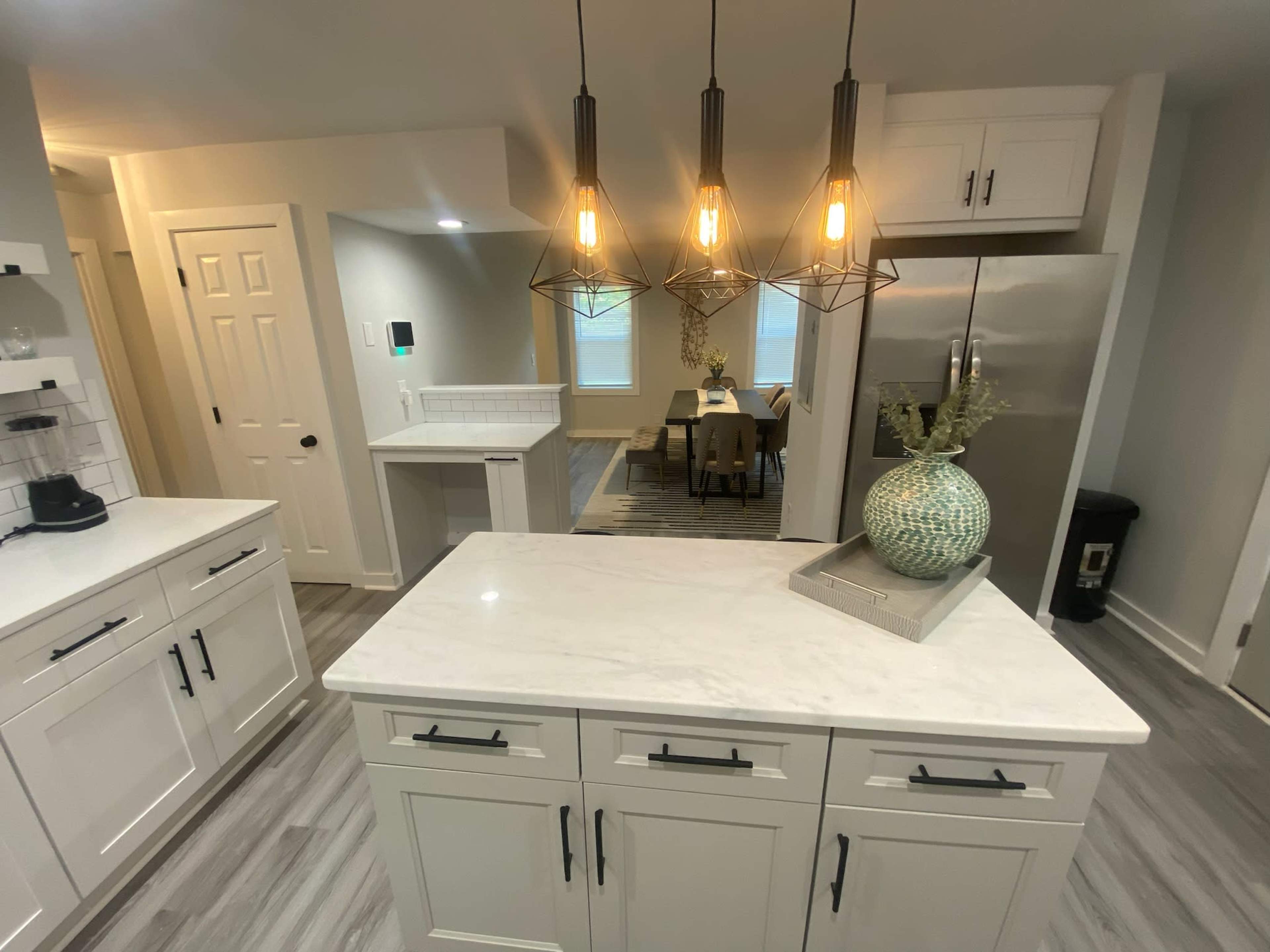 A modern kitchen with white cabinets, a marble island, pendant lighting, and a dining area visible in the background.