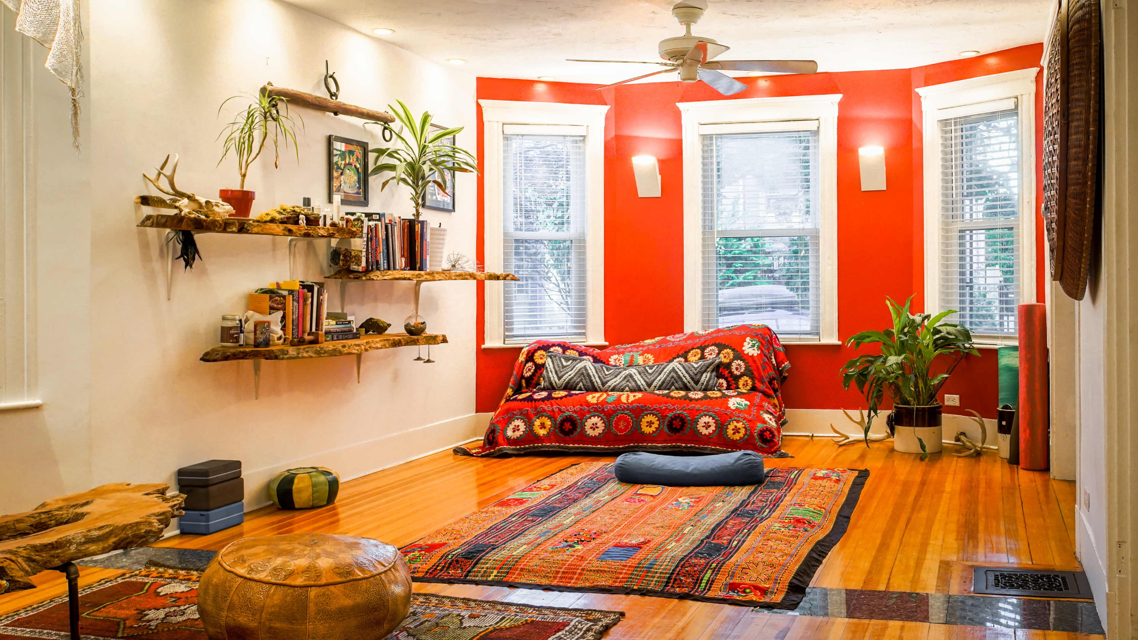 The room features hardwood floors, a red accent wall, a cozy seating area with colorful textiles, bookshelves made from wood, and various plants.
