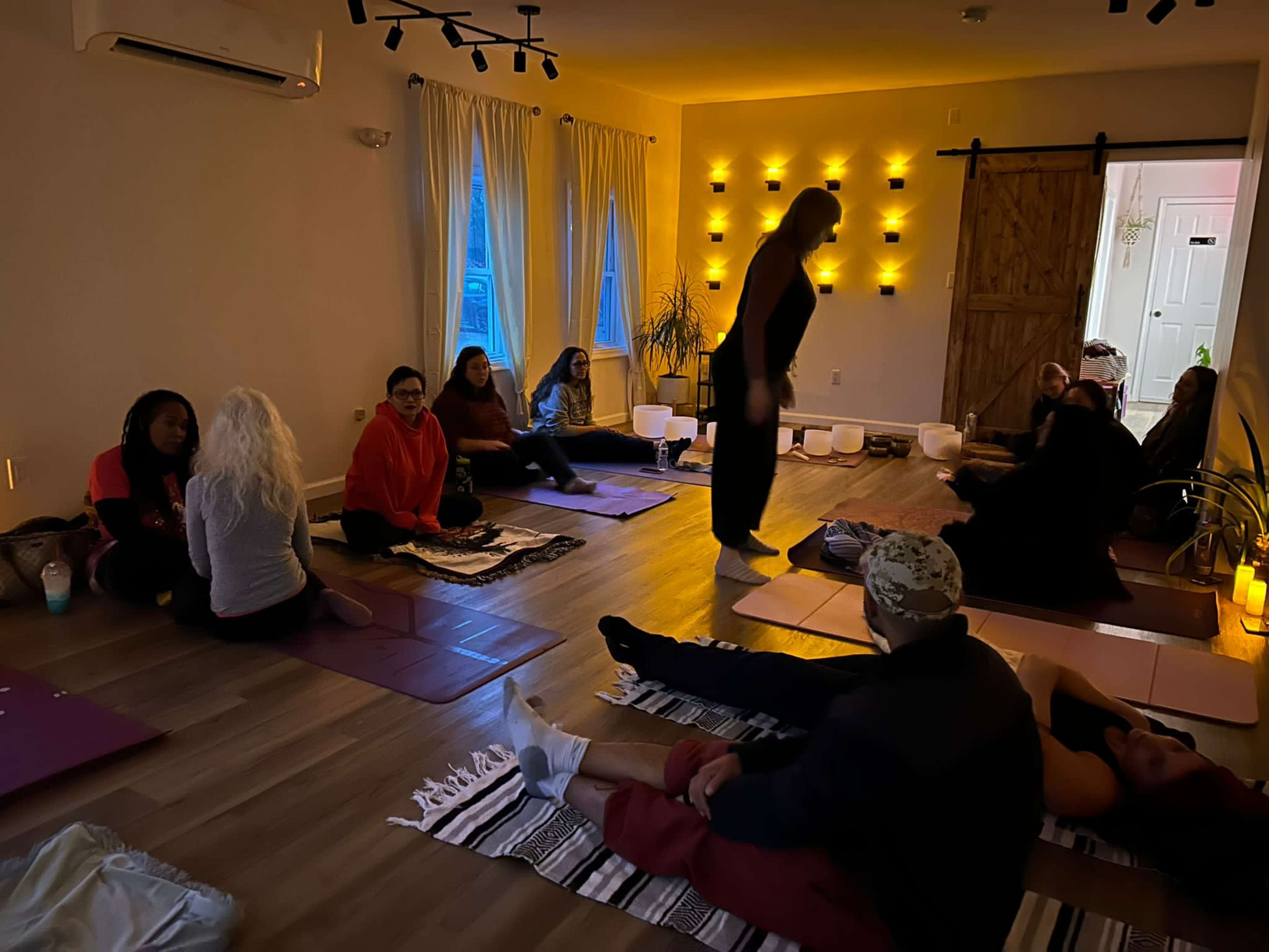 A group of people is seated on mats in a softly lit room, engaging in a relaxation or meditation session.