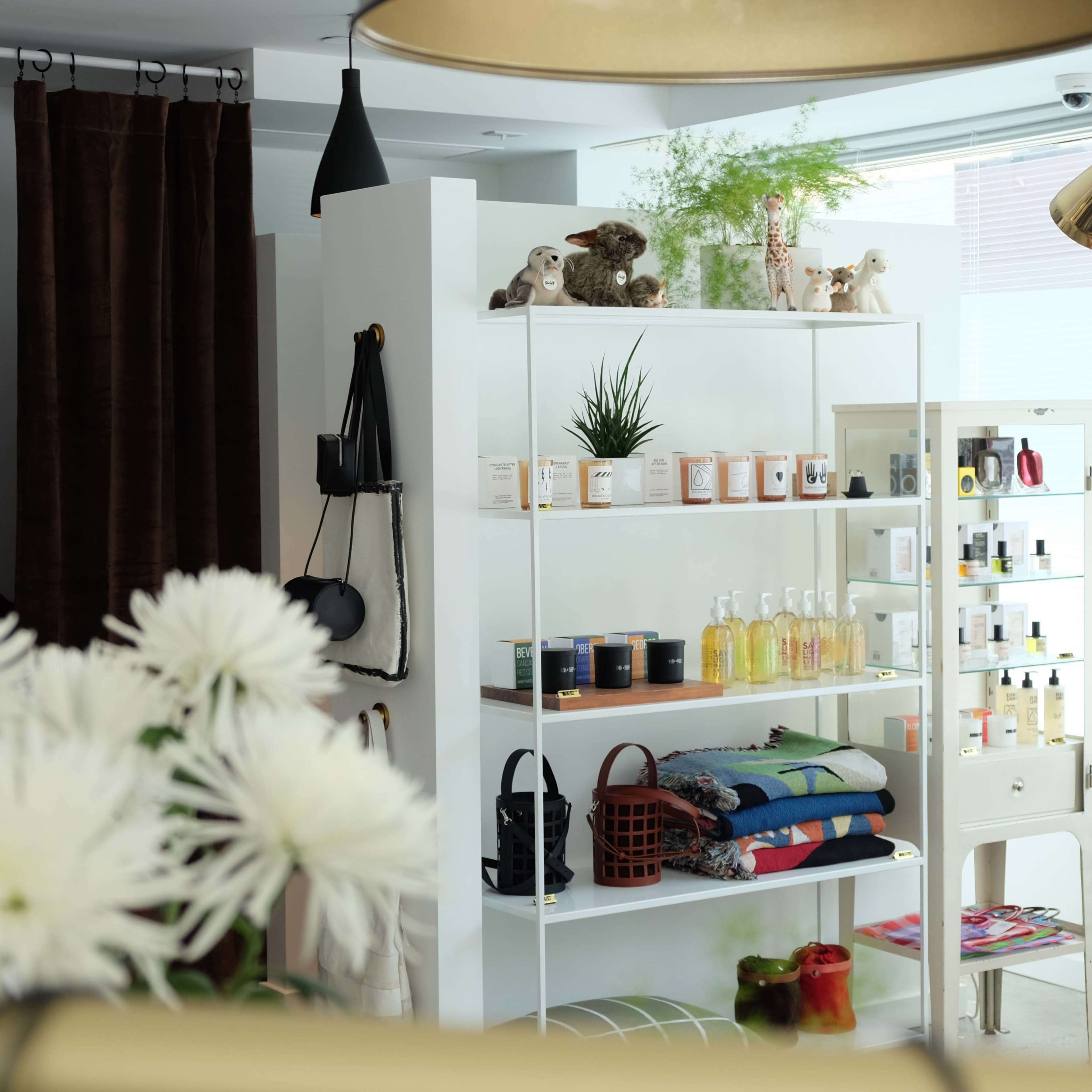 The image shows a modern retail space featuring a white shelving unit stocked with various beauty and home products, alongside decorative items and plants.