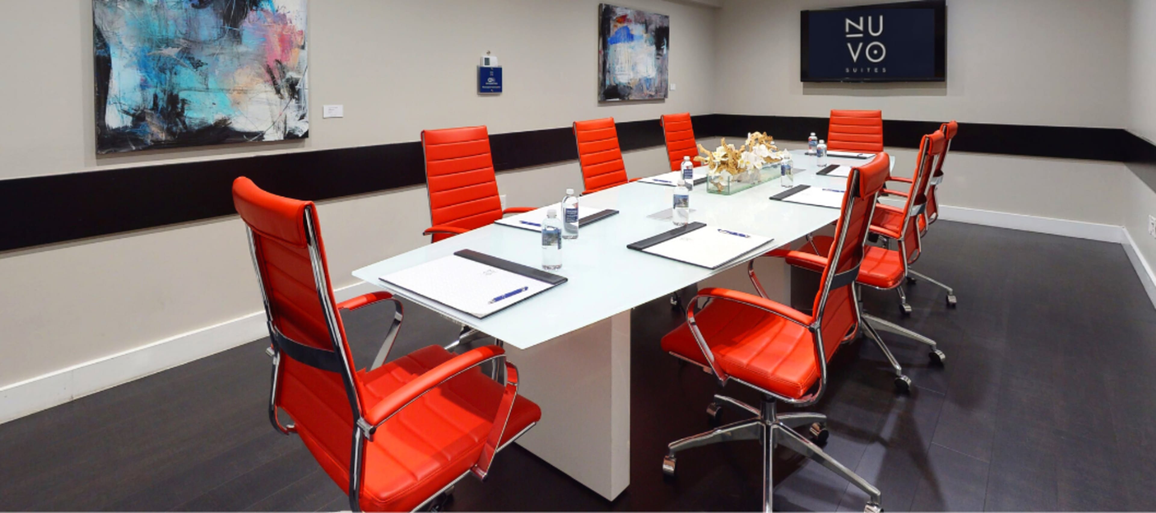 A modern conference room with a long glass table surrounded by red chairs, equipped with notepads and bottles of water.