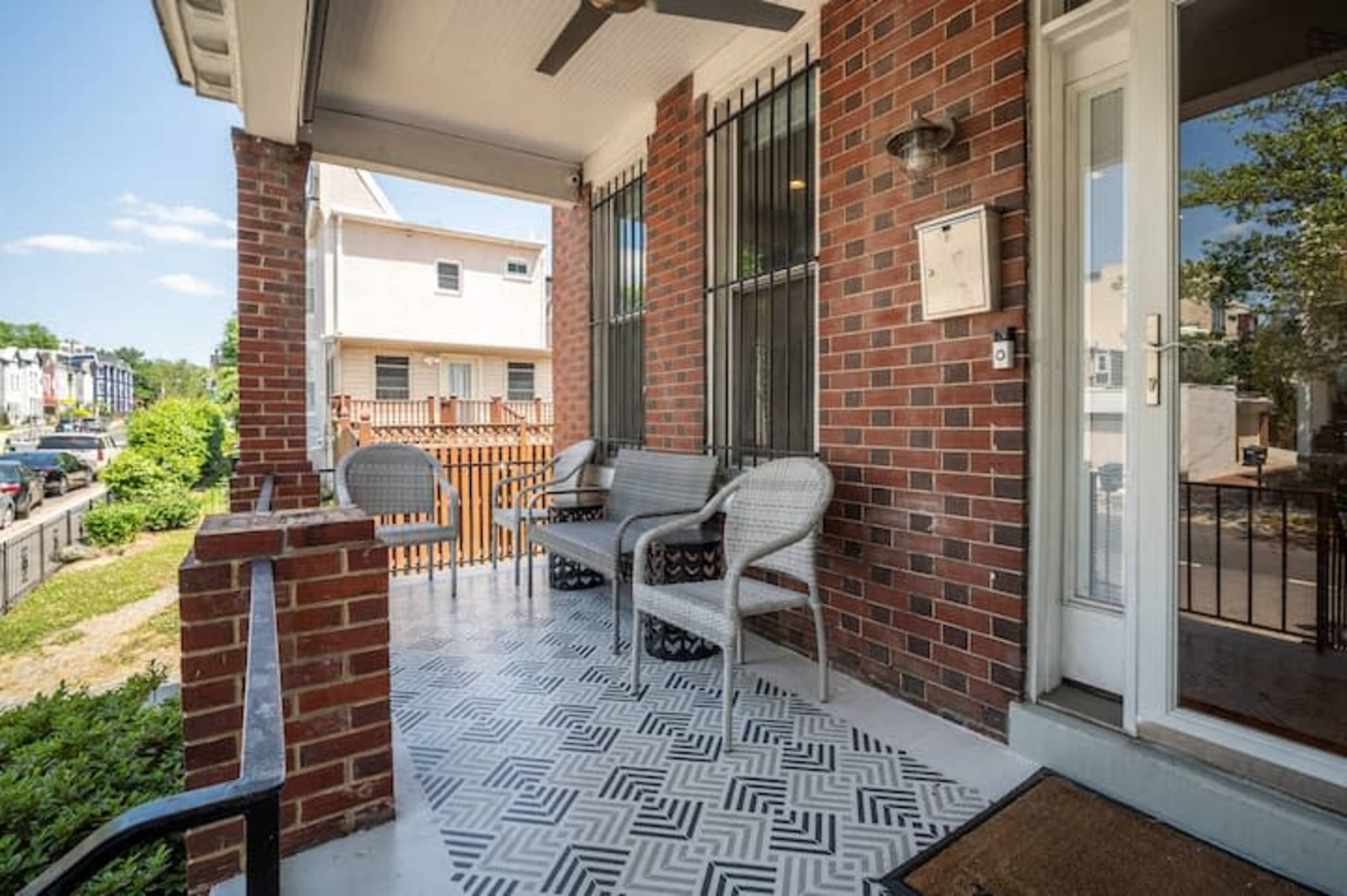 A brick front porch with a patterned tile floor, featuring a seating area of wicker chairs and a small table.