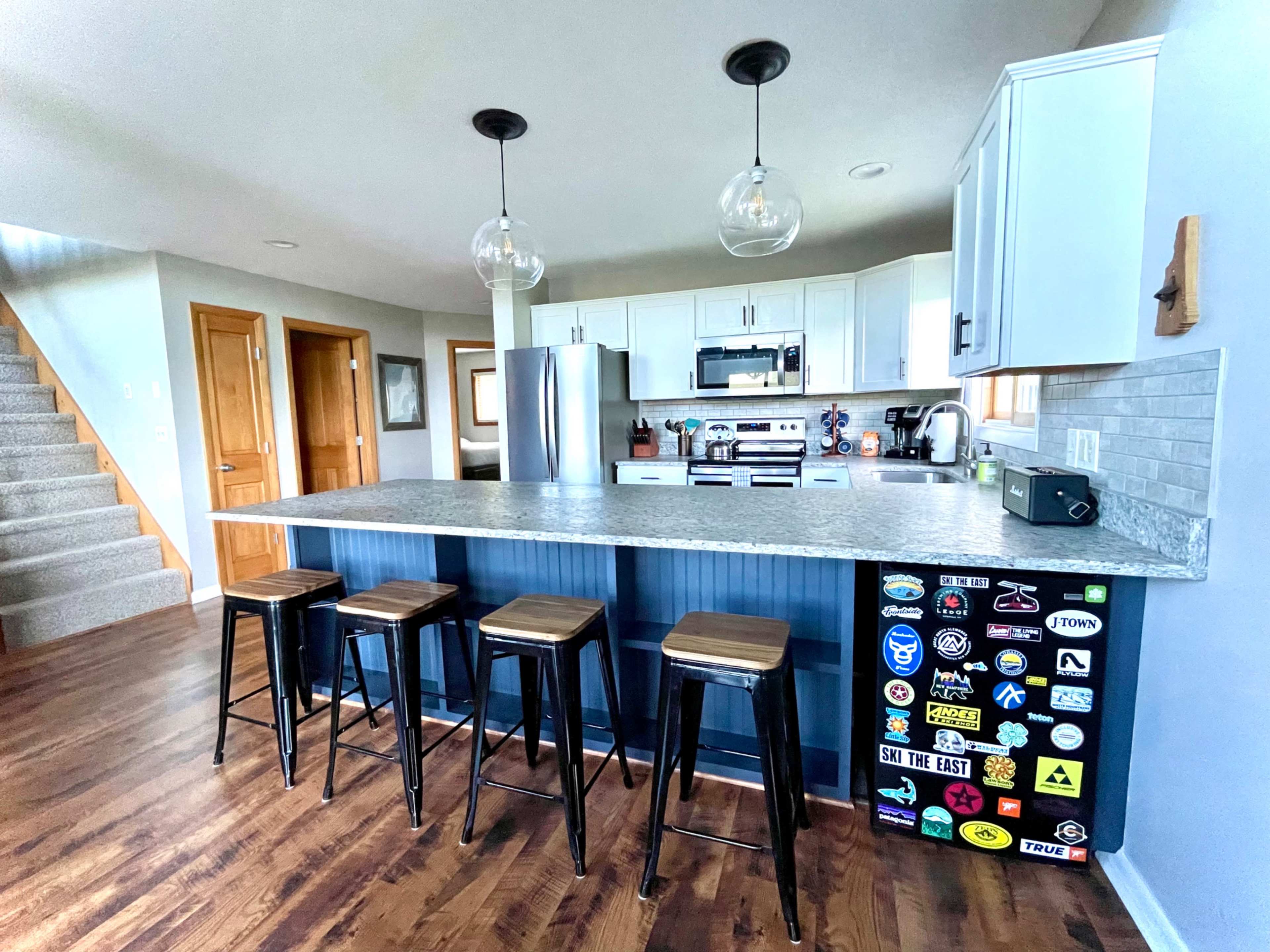 A modern kitchen features a large island with four black stools, stainless steel appliances, and a mix of blue and white cabinetry.