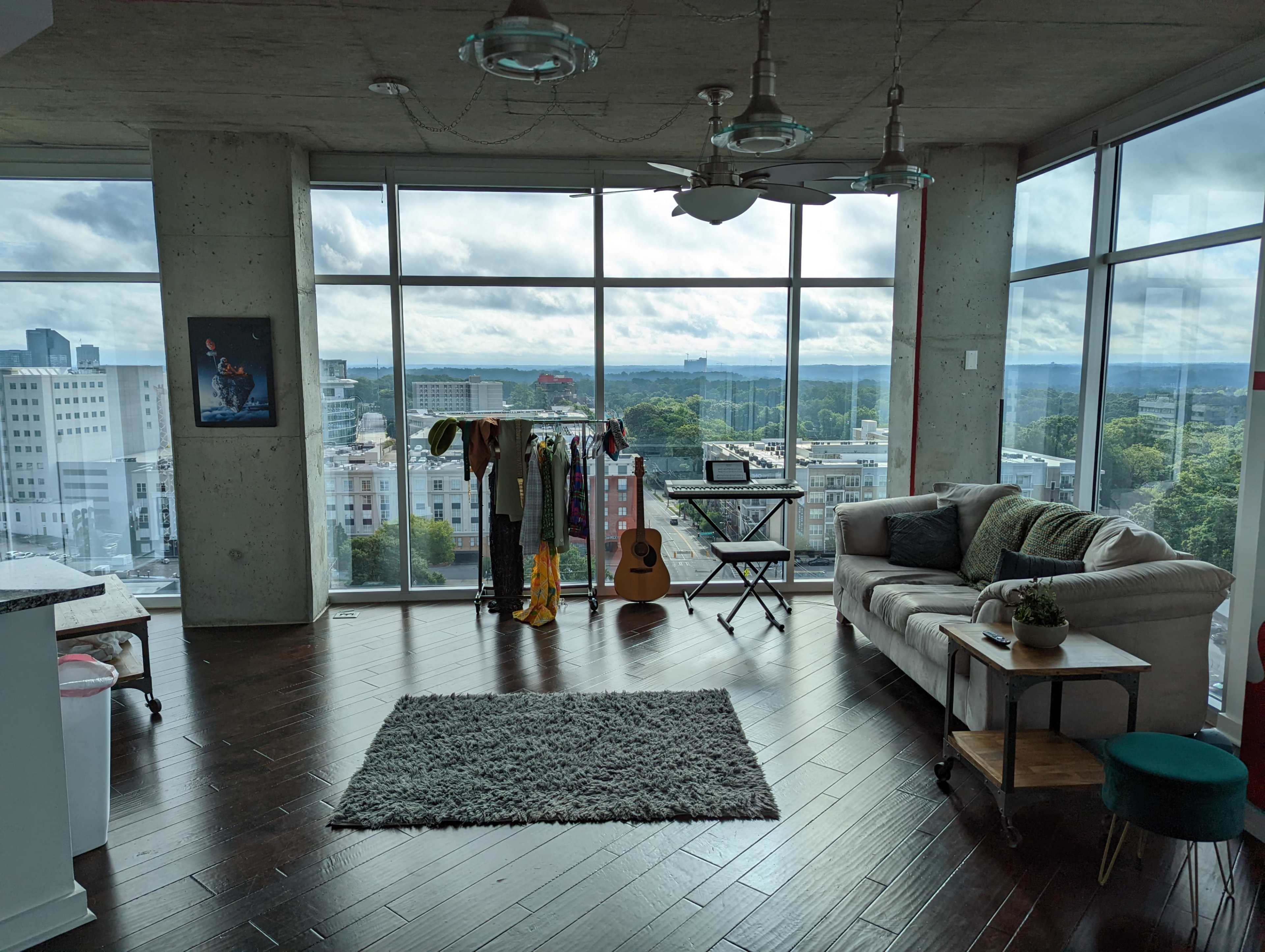 A modern loft features large windows with a city view, a sofa, a clothing rack, a guitar, and a rug on wooden flooring.