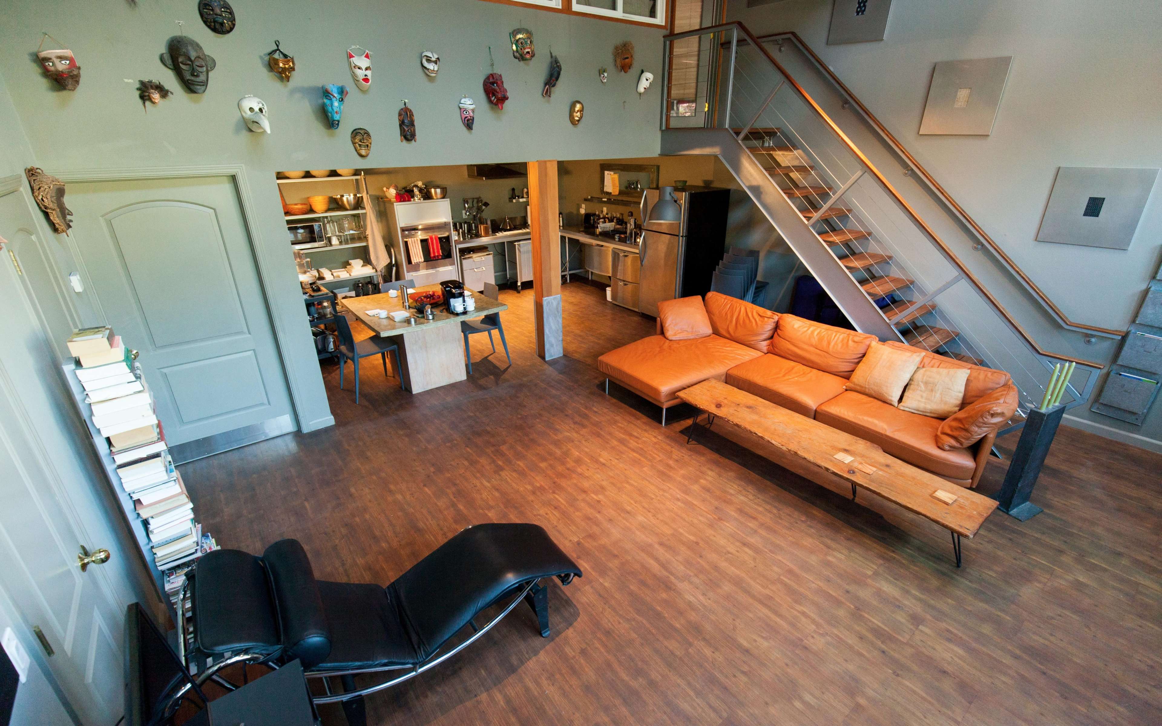A spacious, open-plan living area featuring a brown leather sofa, a wooden coffee table, a staircase, and a kitchen with various appliances, along with decorative masks on the wall.