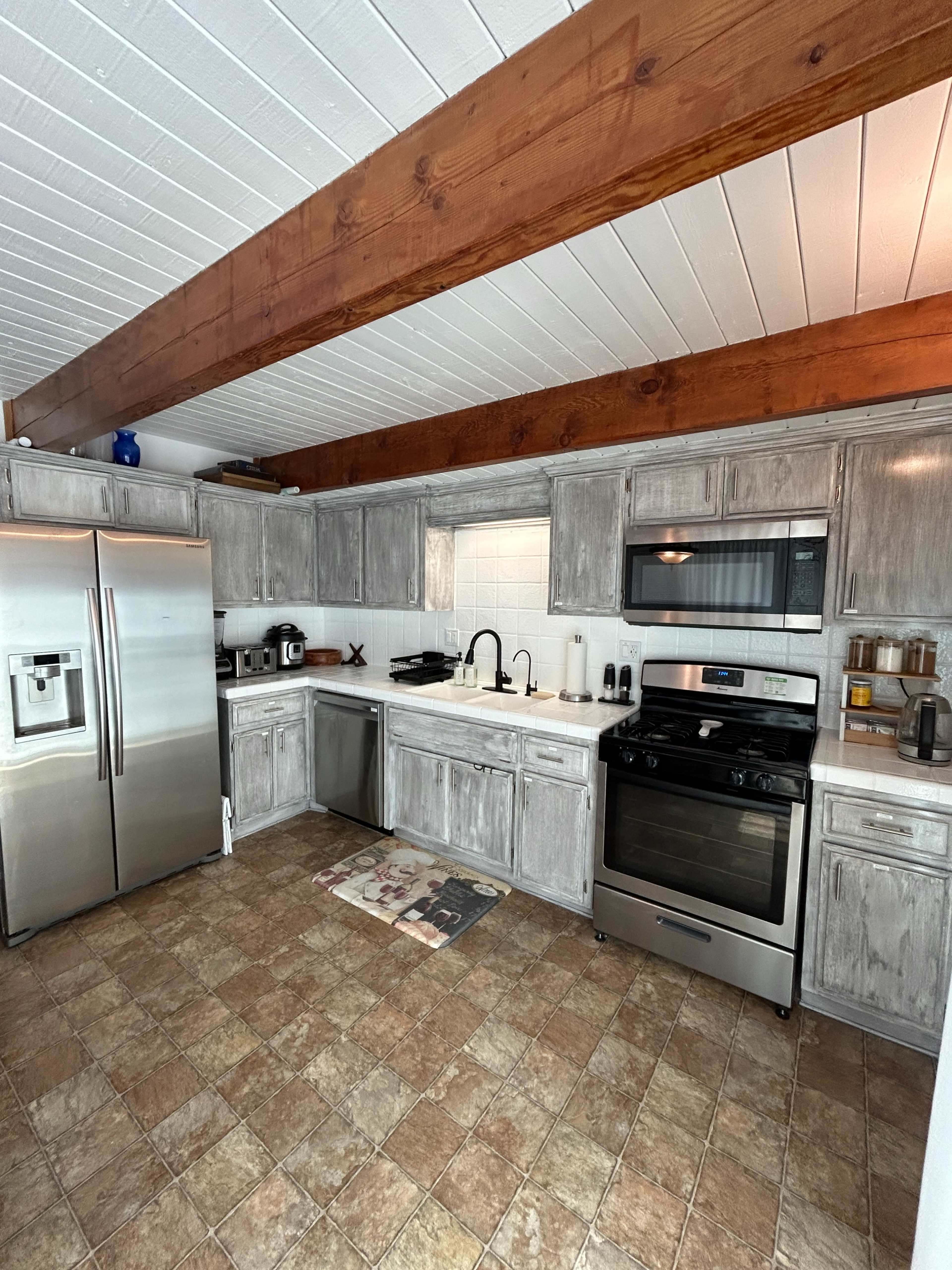 The kitchen features stainless steel appliances, wooden beams on the ceiling, and light gray cabinetry with a tile floor.