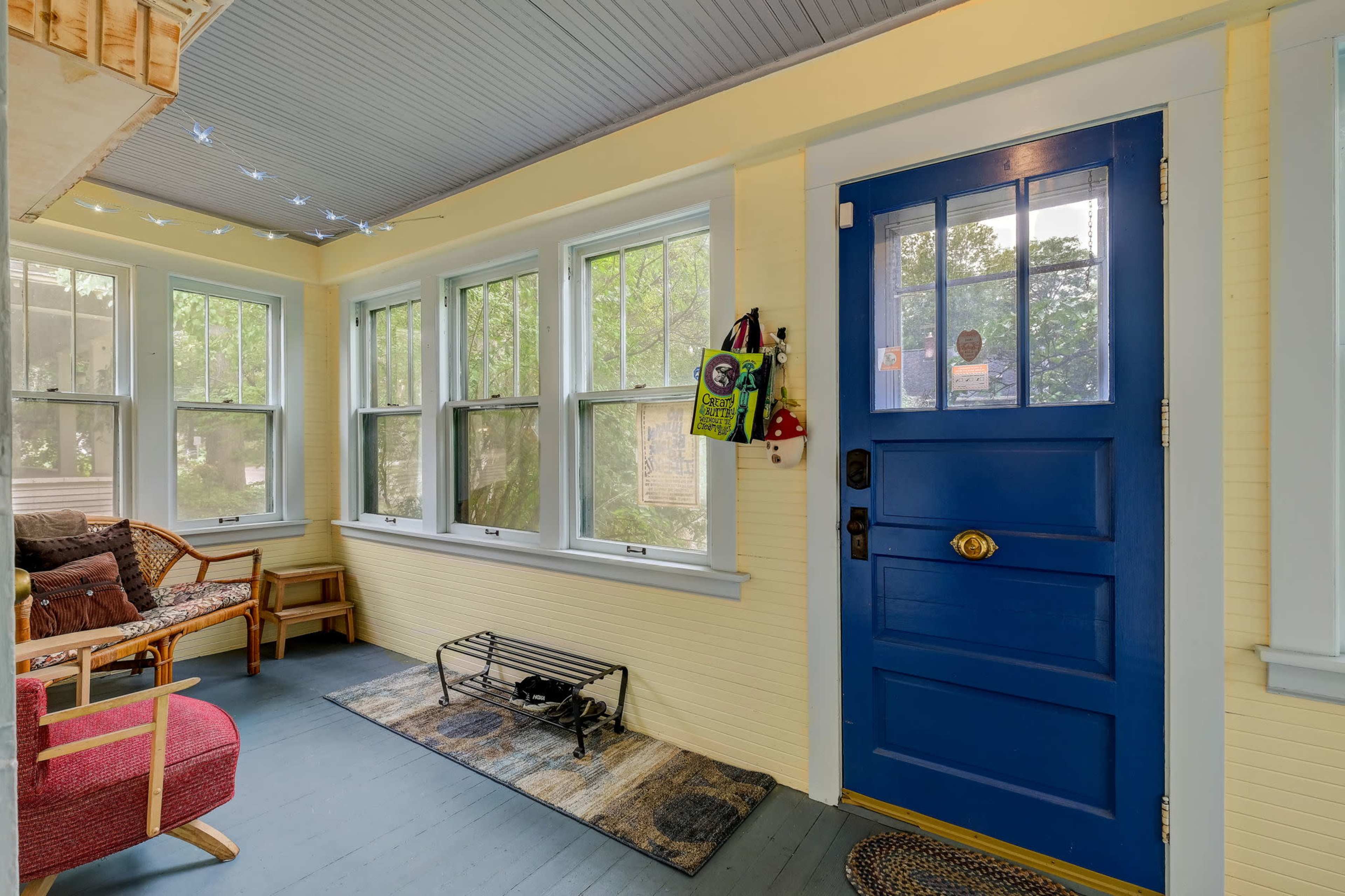 A bright sunroom with large windows, a blue front door, and a seating area featuring a wicker chair and a small rug.