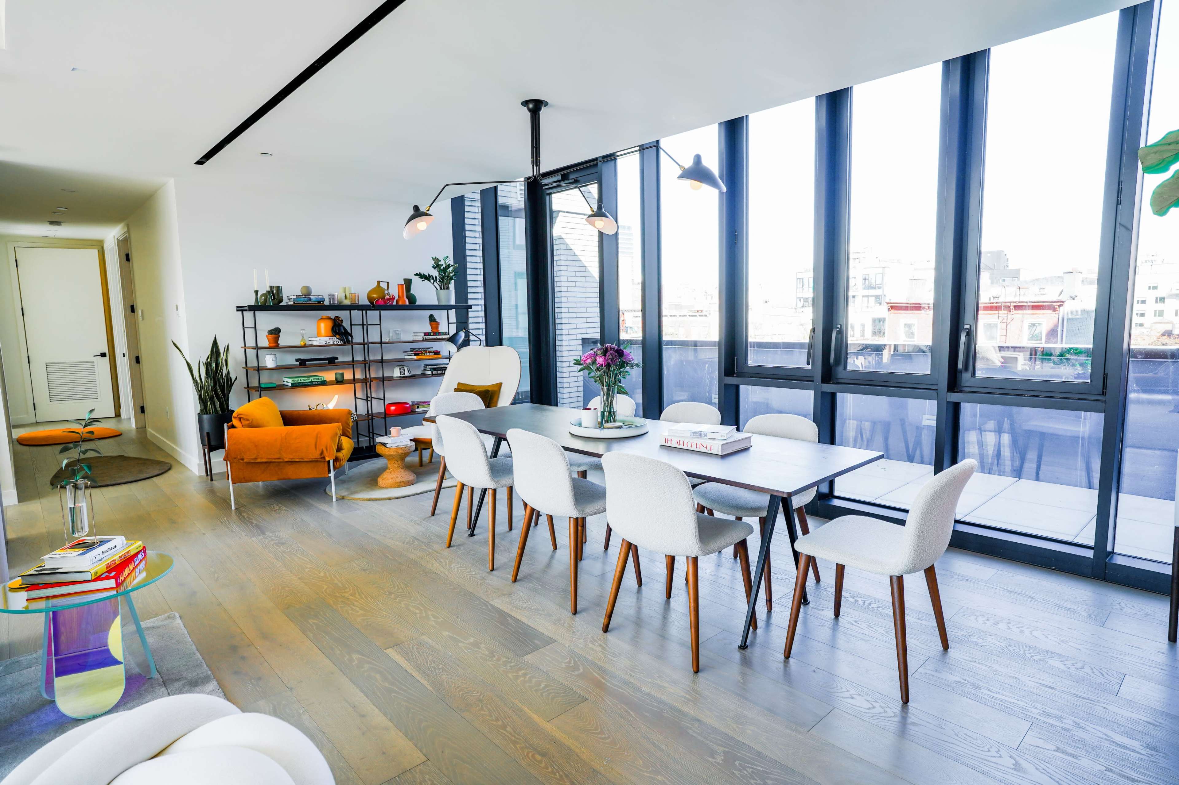 A modern dining area with a table surrounded by white chairs, and large windows letting in natural light, overlooking a cityscape.