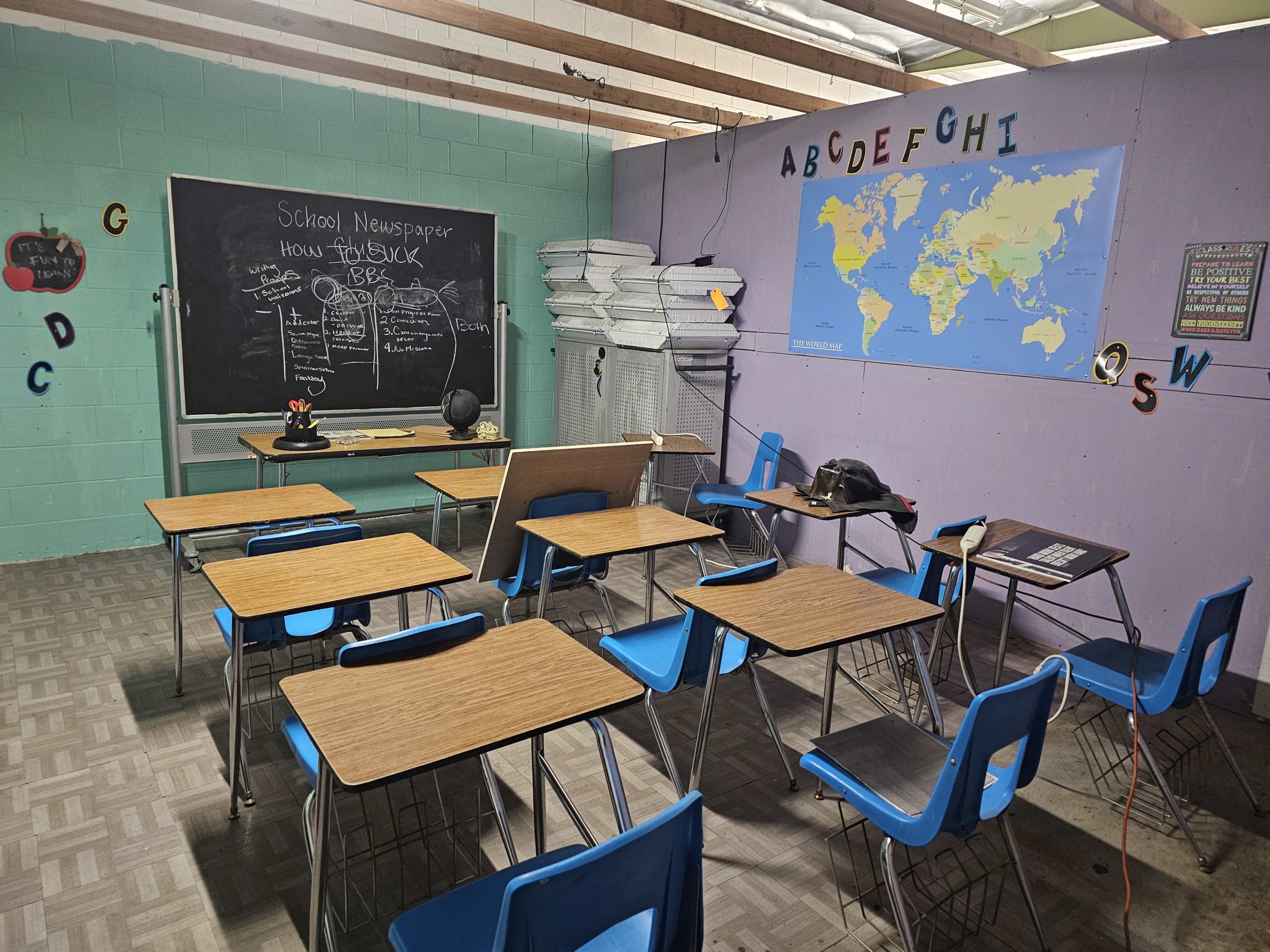 The image shows a classroom with desks arranged in rows, a blackboard displaying a newspaper topic, stacks of paper in the corner, and a world map on the wall.