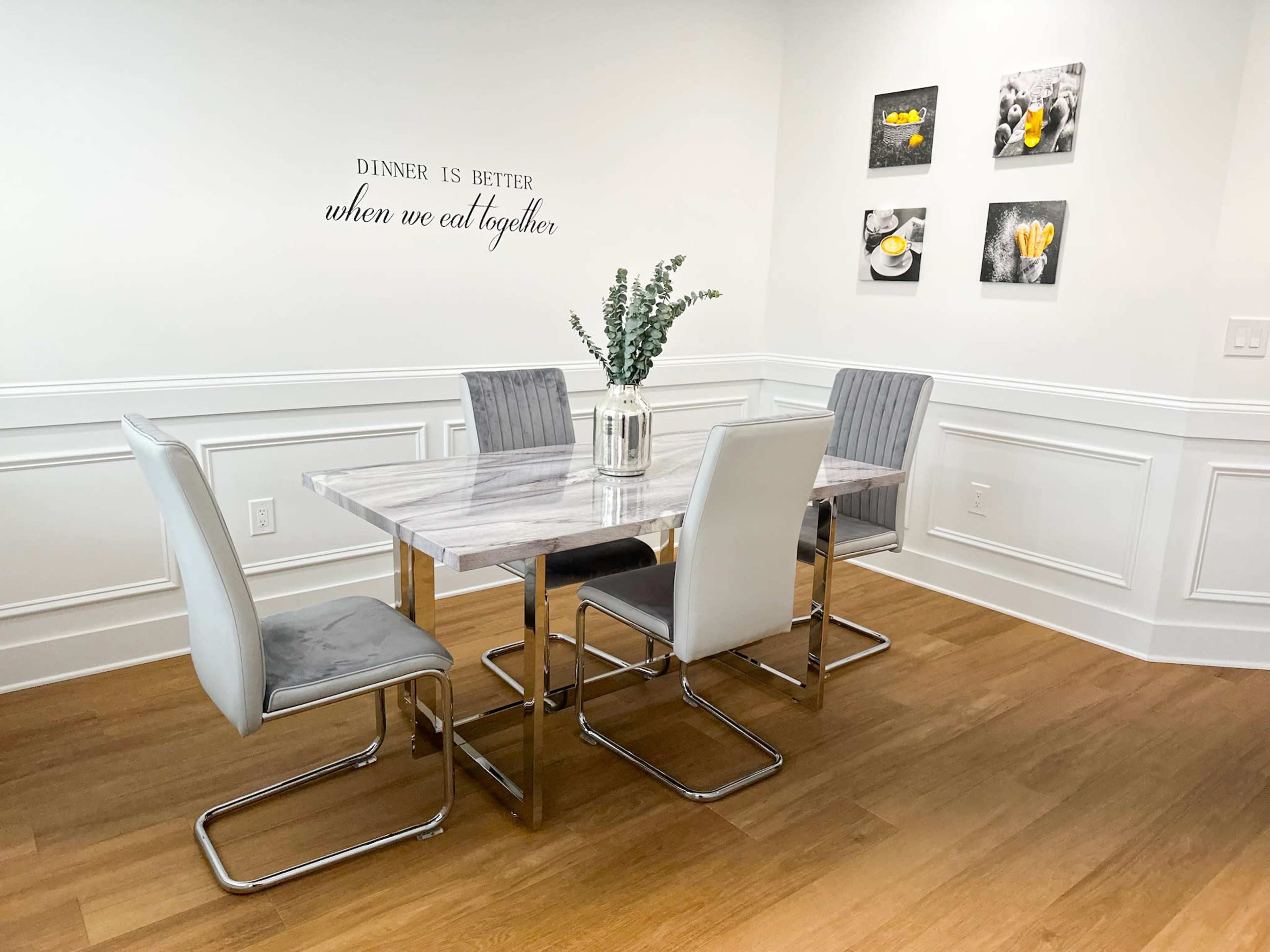 A dining table with four chairs is set against a light-colored wall featuring decorative pictures and a quote about dining together.