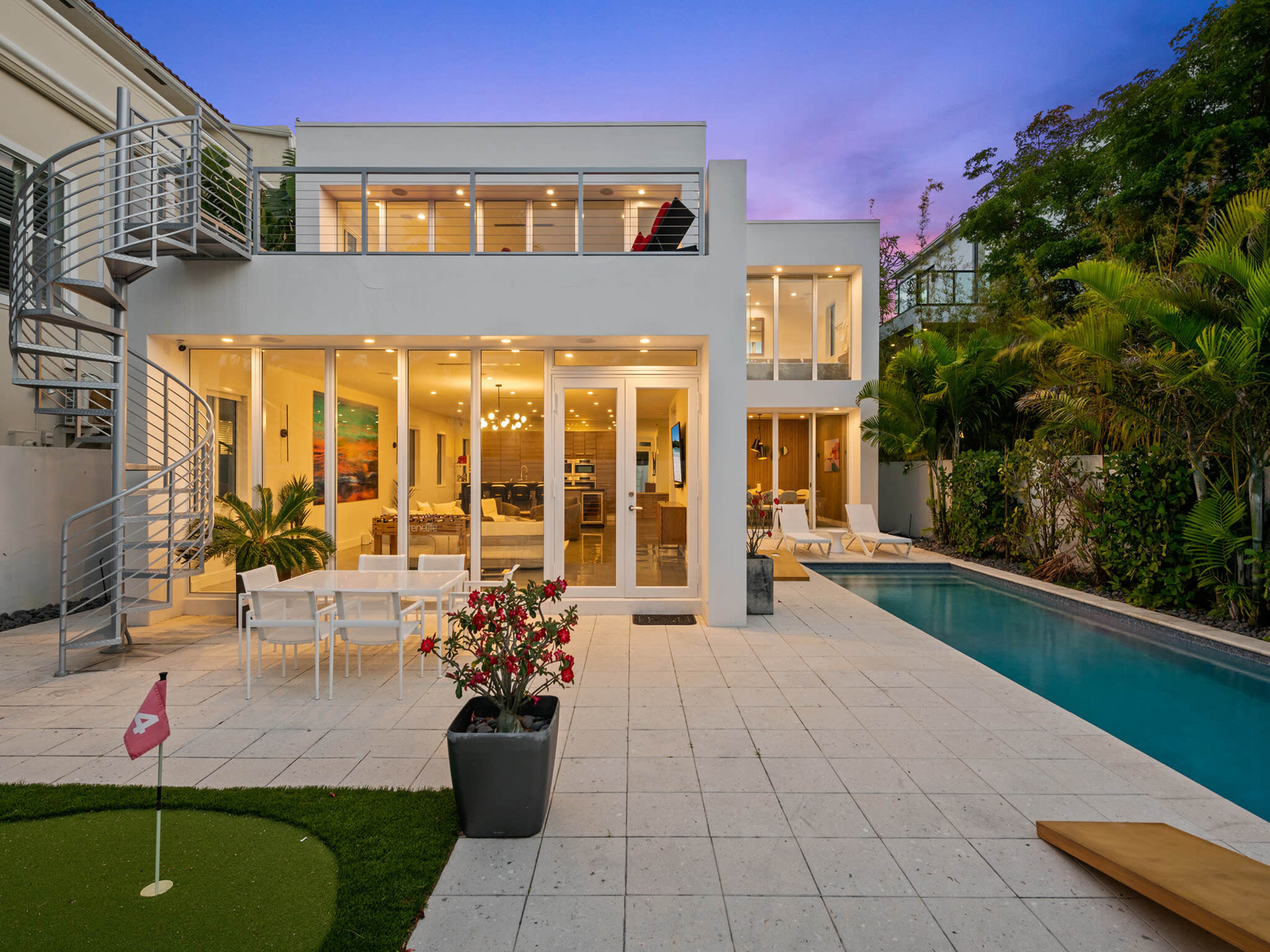 The image shows a modern house with large glass windows, a swimming pool, and a small golf putting area surrounded by tropical plants.