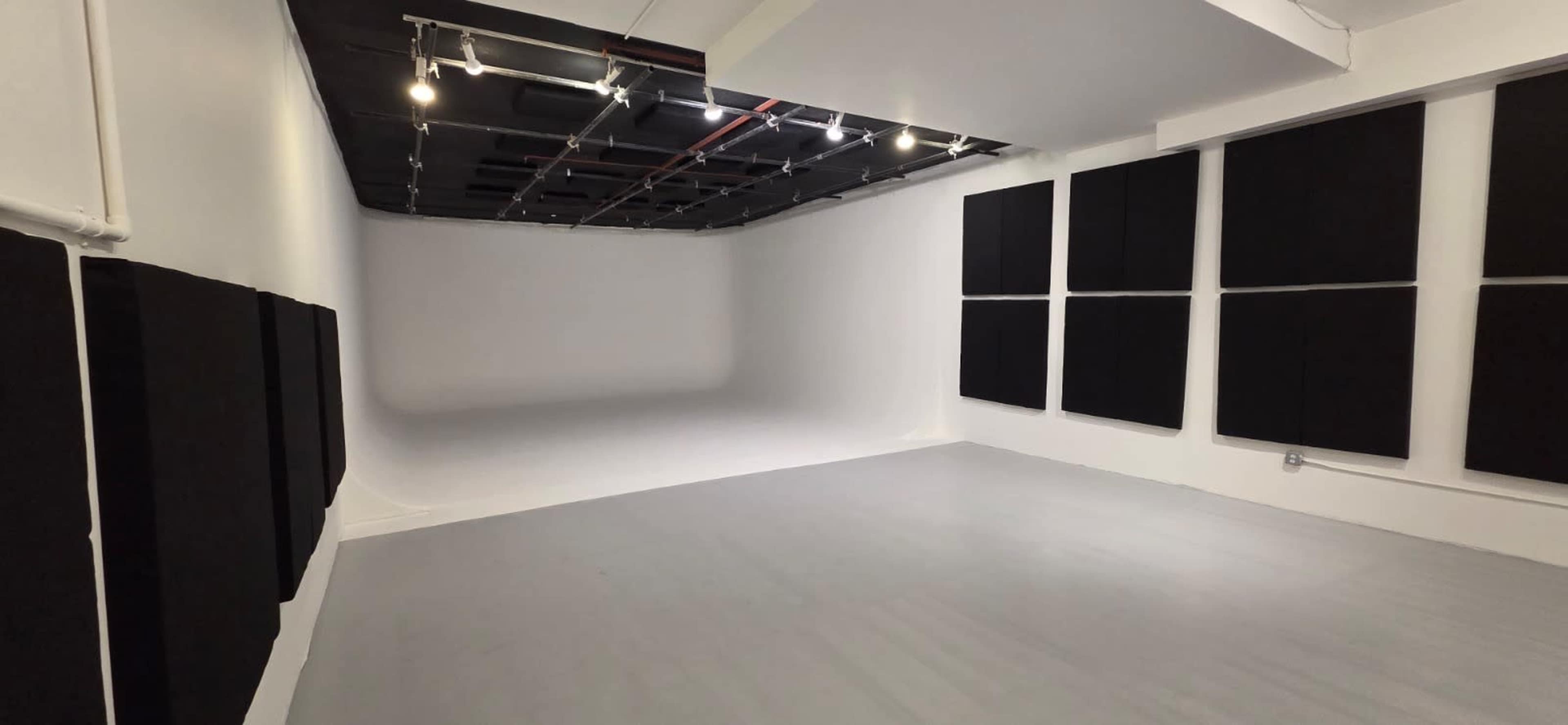 The image shows a spacious photography studio with a light gray floor, white walls, and several black acoustic panels mounted on the walls.