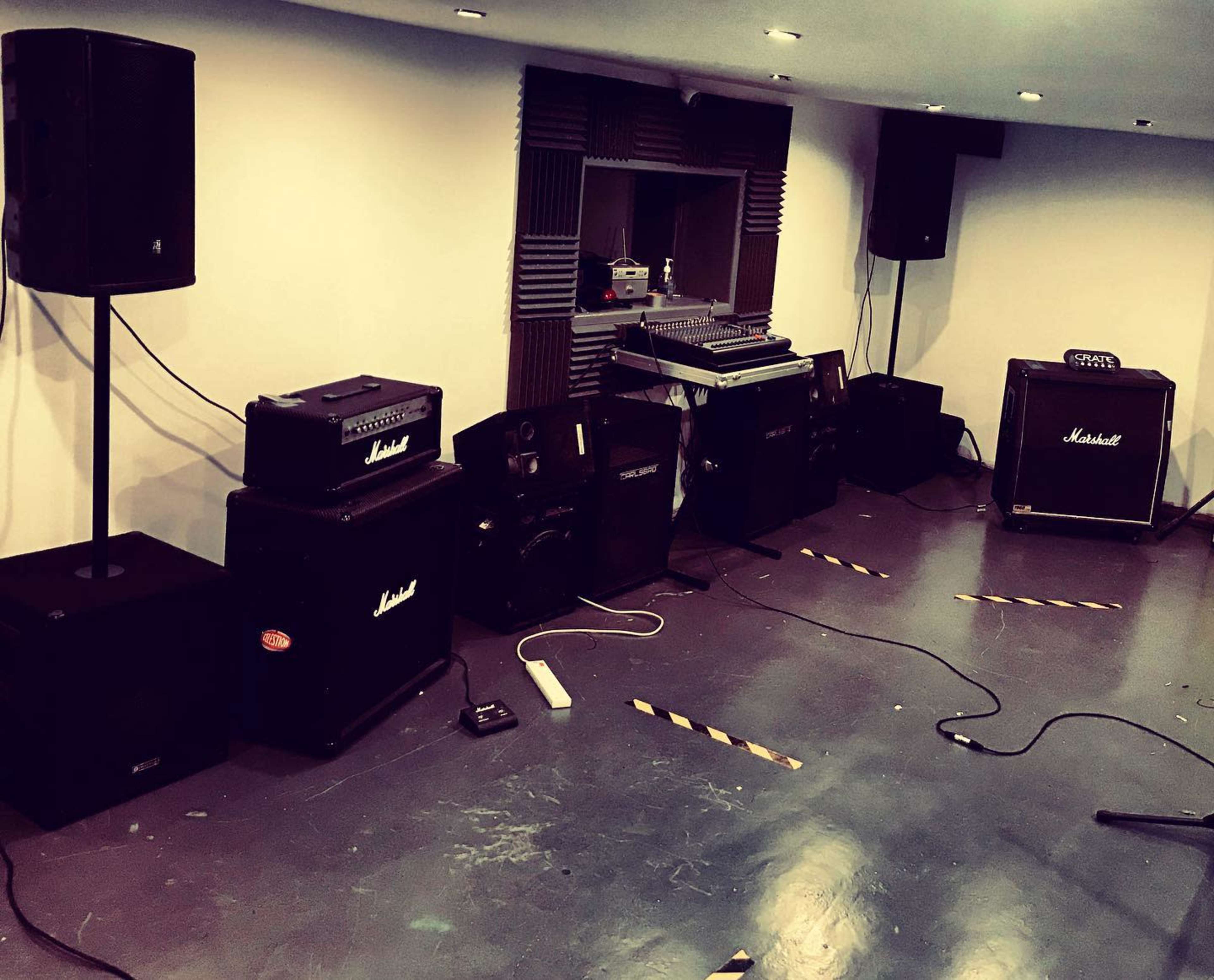 The image shows a music rehearsal space with multiple amplifiers, a mixing console, and audio equipment arranged along the walls.