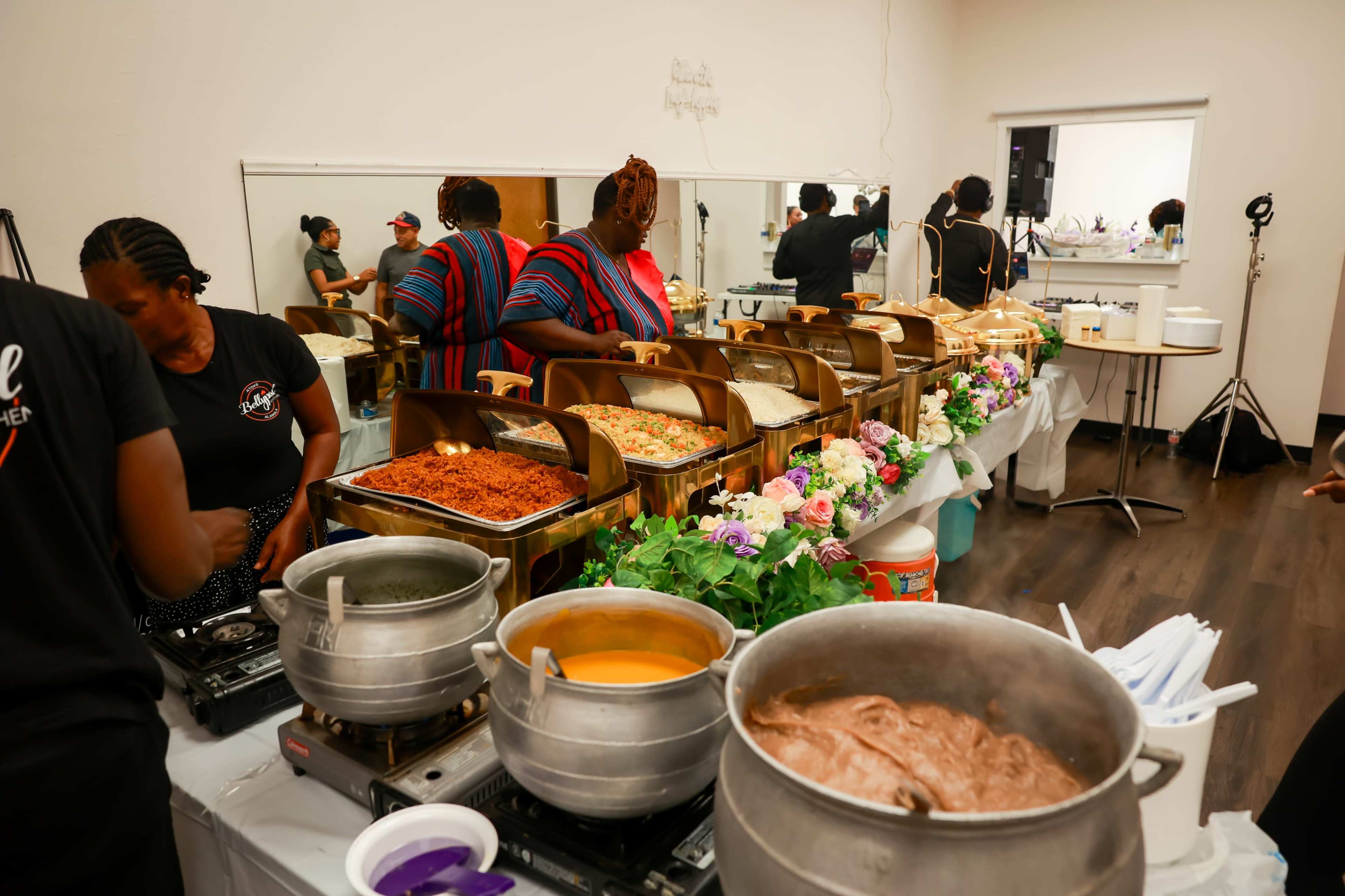 A buffet table is set up with various dishes in chafing dishes and pots, while people are seen preparing food in a kitchen area.