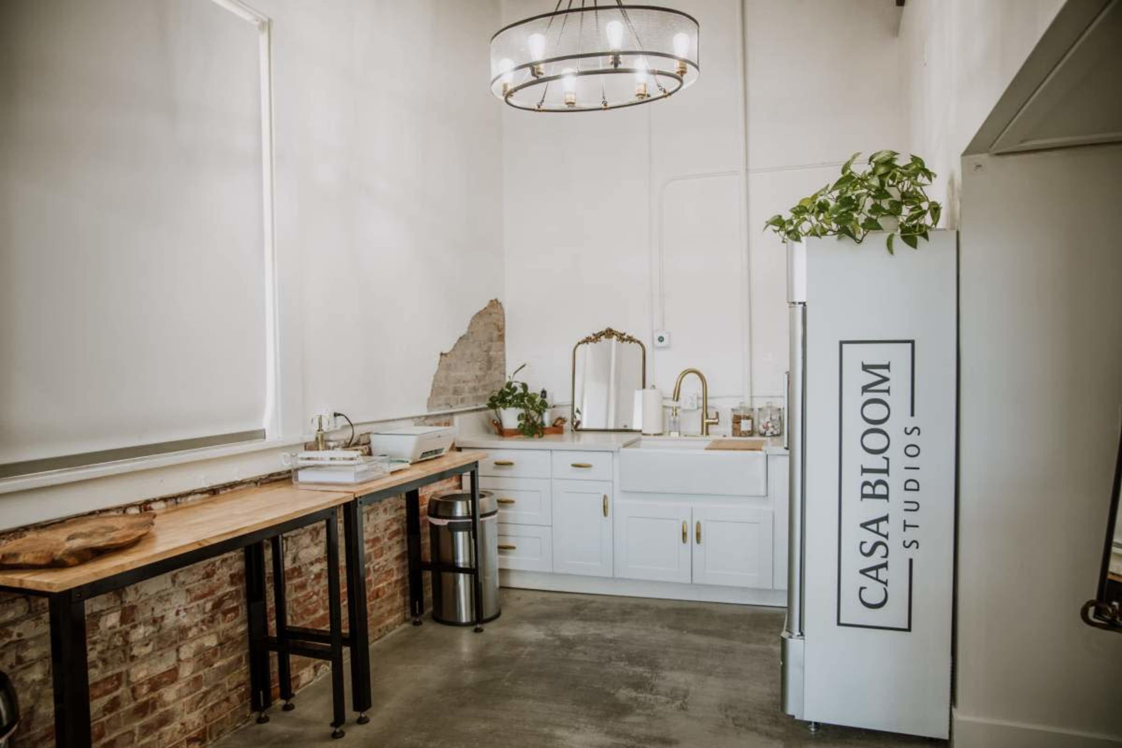 The image shows a modern studio kitchen with white cabinetry, a wooden table, and a large refrigerator labeled "CASA BLOOM STUDIOS."