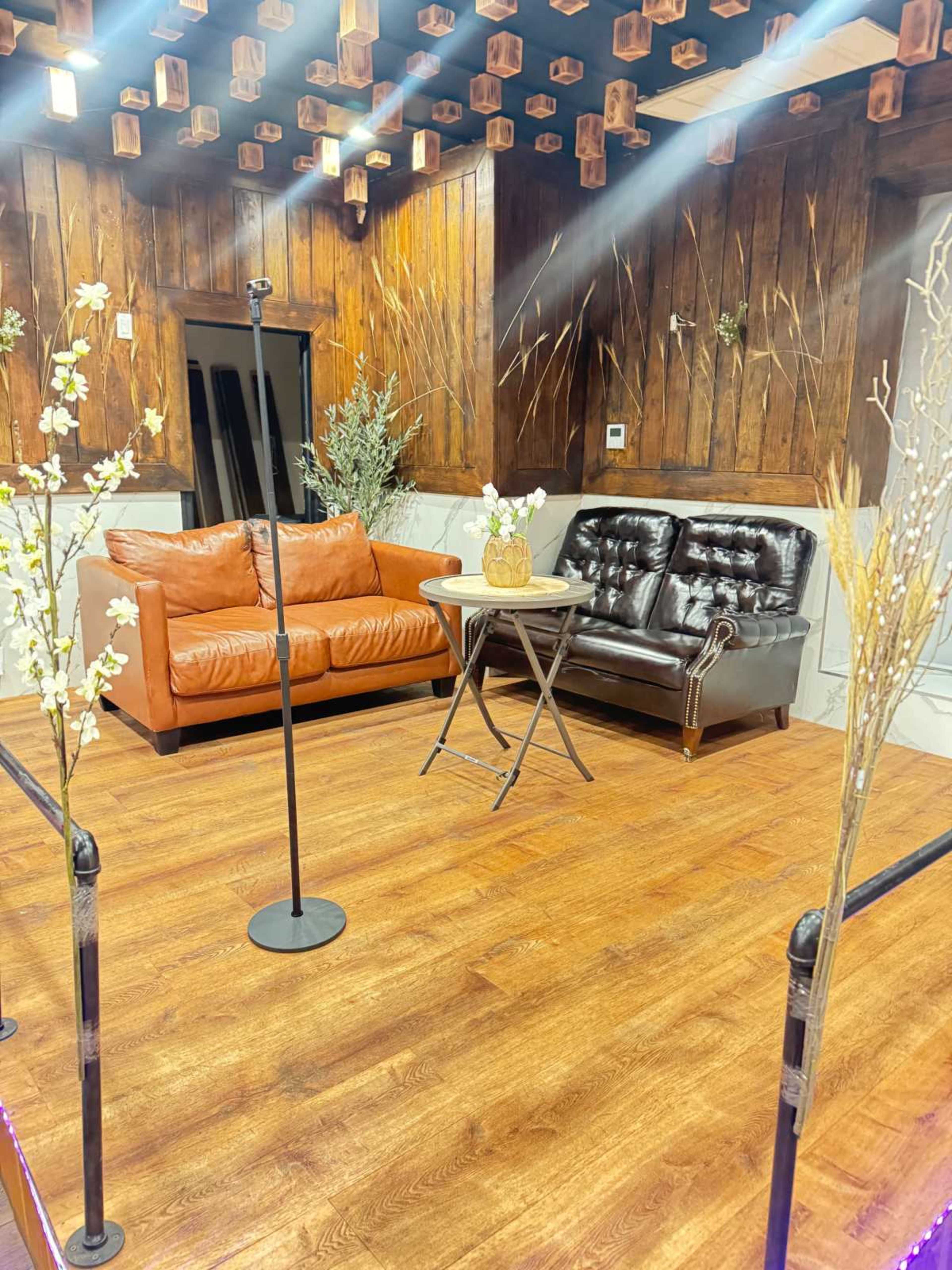The image shows a cozy performance space featuring two sofas, a small round table, decorative plants, and wooden wall panels.