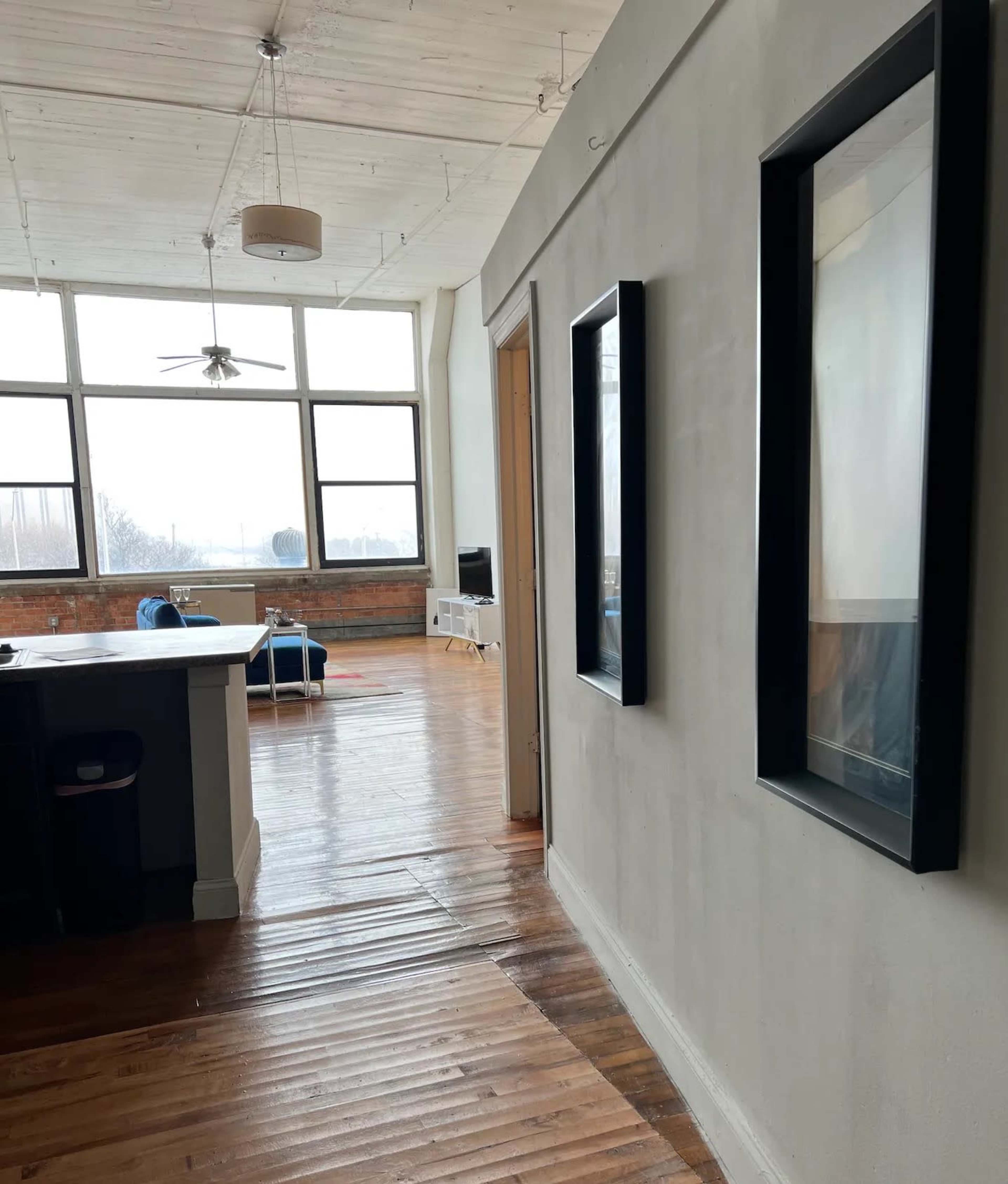 The image shows a hallway with two framed pictures on the gray wall, leading to a spacious room with large windows and wooden flooring.