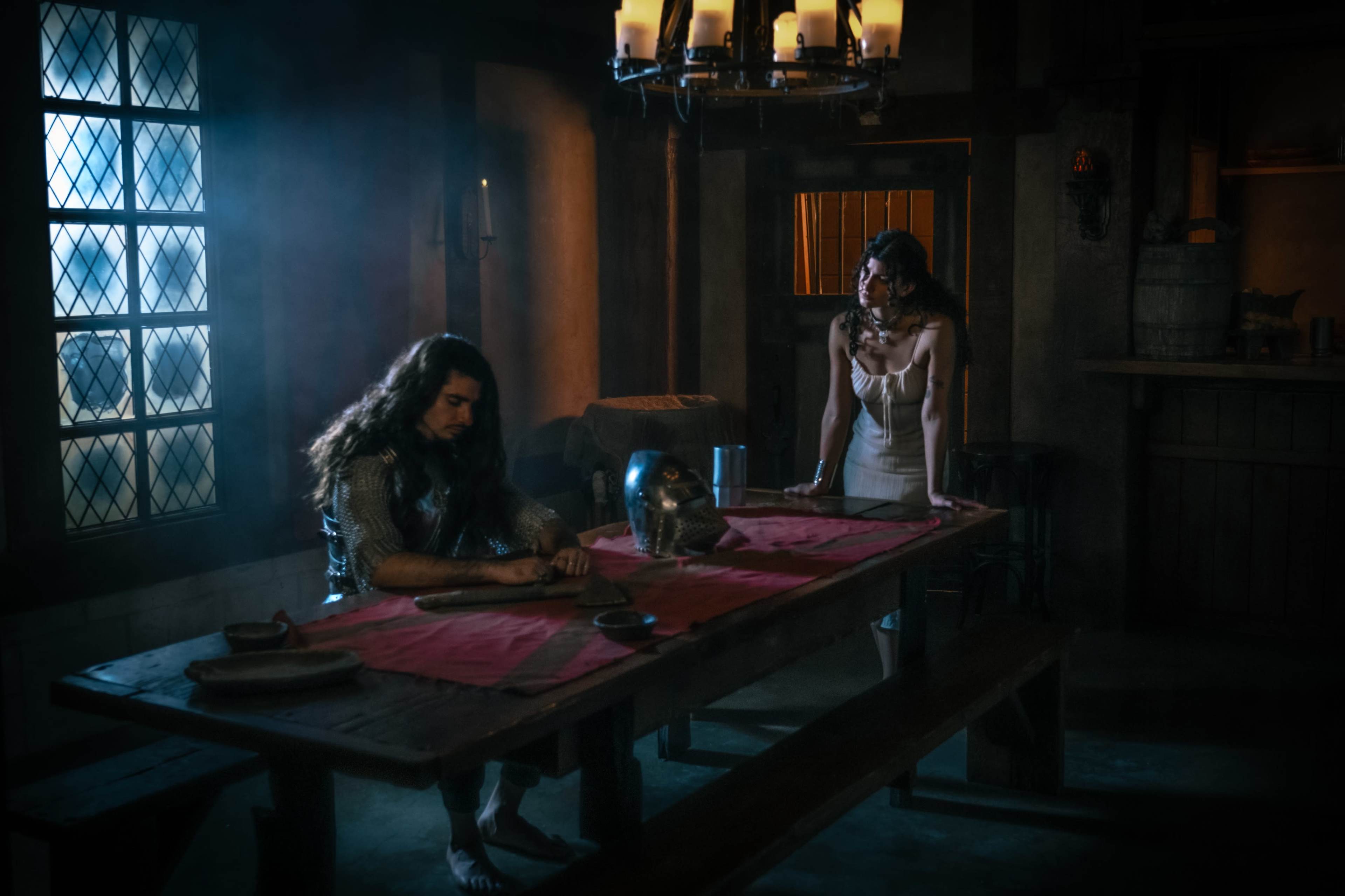 A man with long hair sits at a wooden table in a dimly lit room while a woman stands nearby, both surrounded by medieval decor.