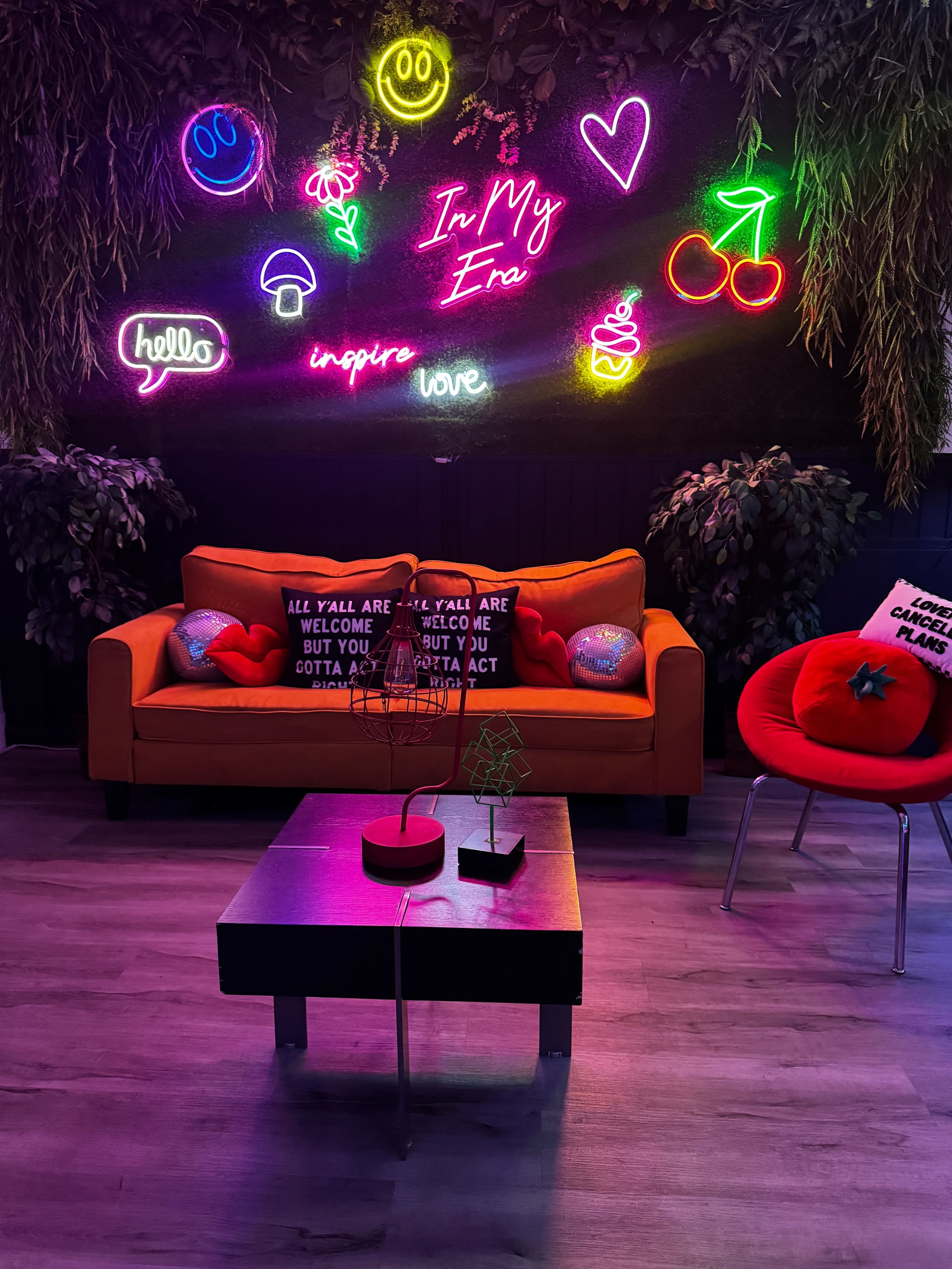The image shows a colorful modern lounge area featuring a bright orange sofa, a round red chair, and a table, all set against a wall adorned with neon signs and decorative plants.