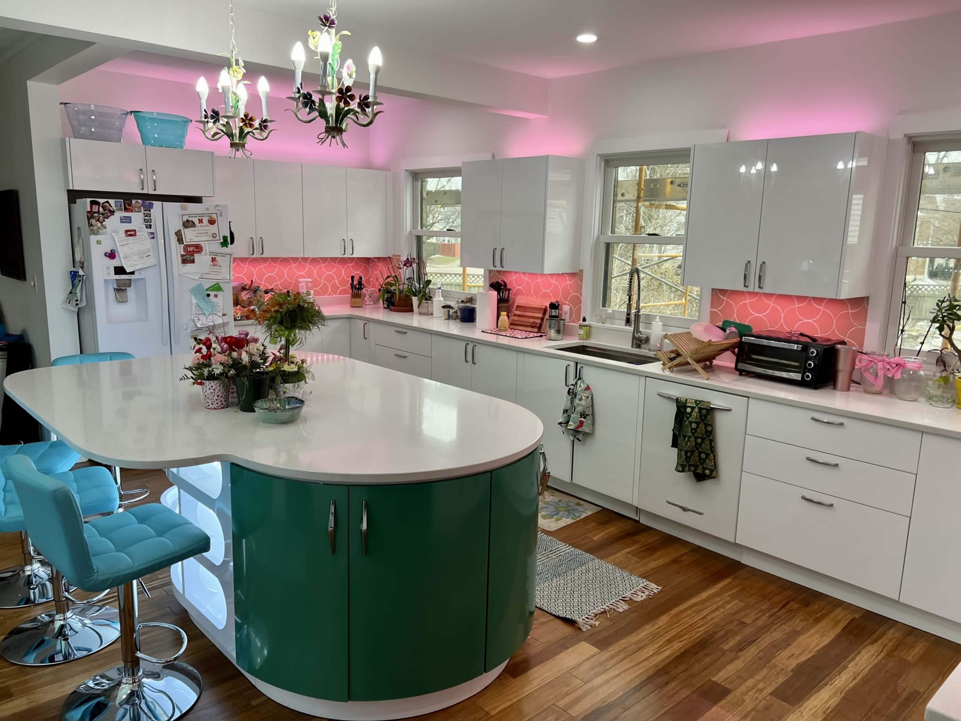 A modern kitchen with a large central island in teal, colorful LED lighting, and a variety of appliances and decorations along the countertops.