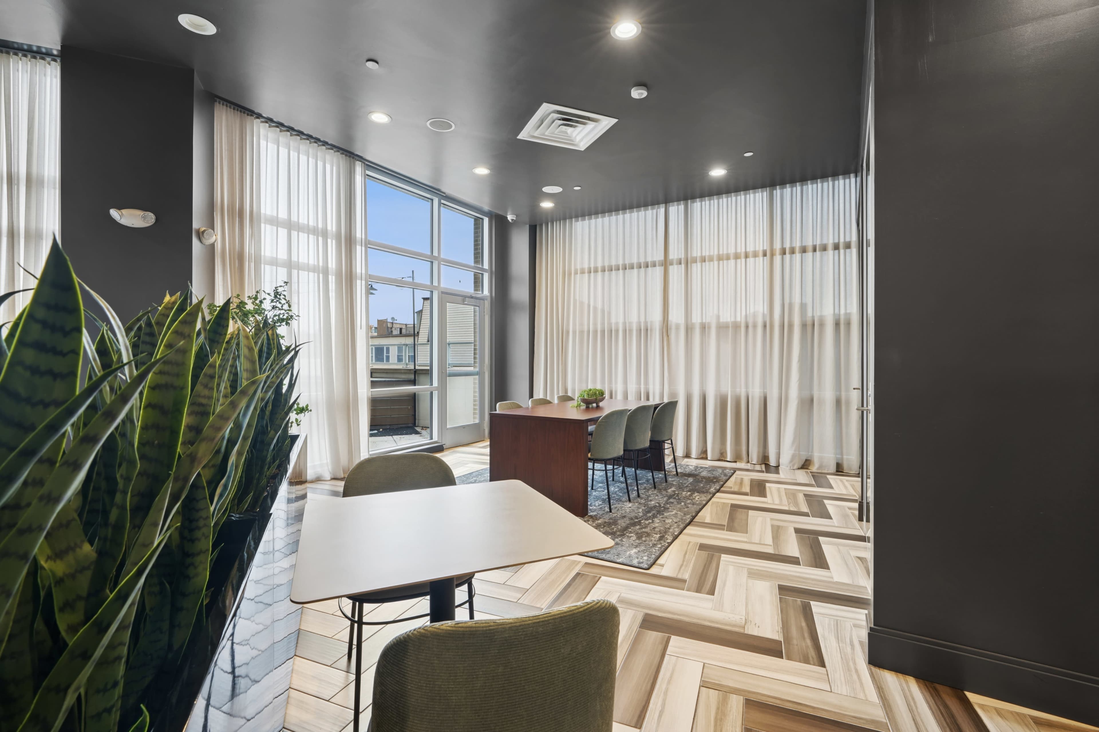 The image features a modern interior space with large windows, a wooden table surrounded by chairs, and indoor plants near the entrance.