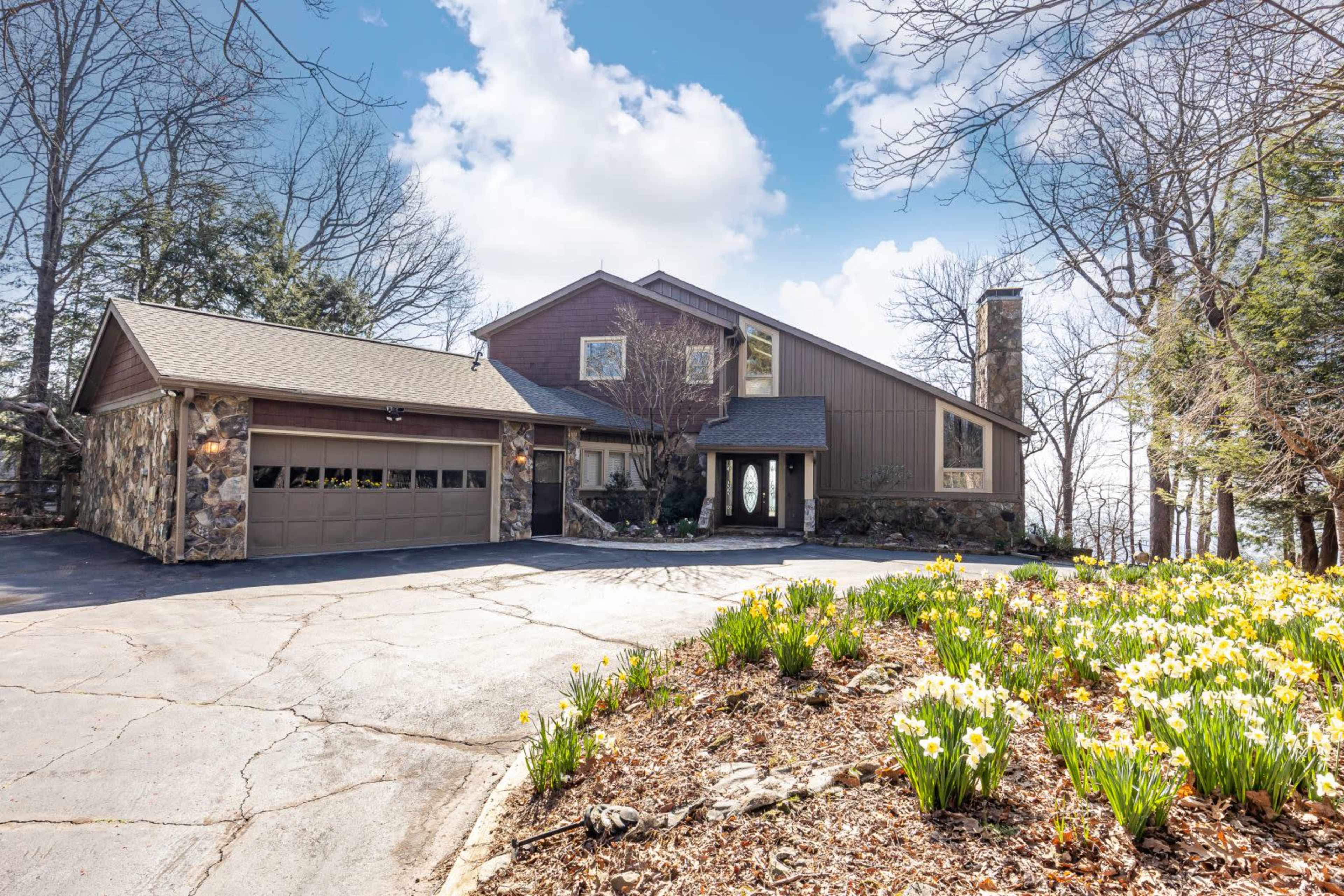 A modern, two-story house with a stone facade sits beside a paved driveway lined with blooming daffodils and surrounded by trees.
