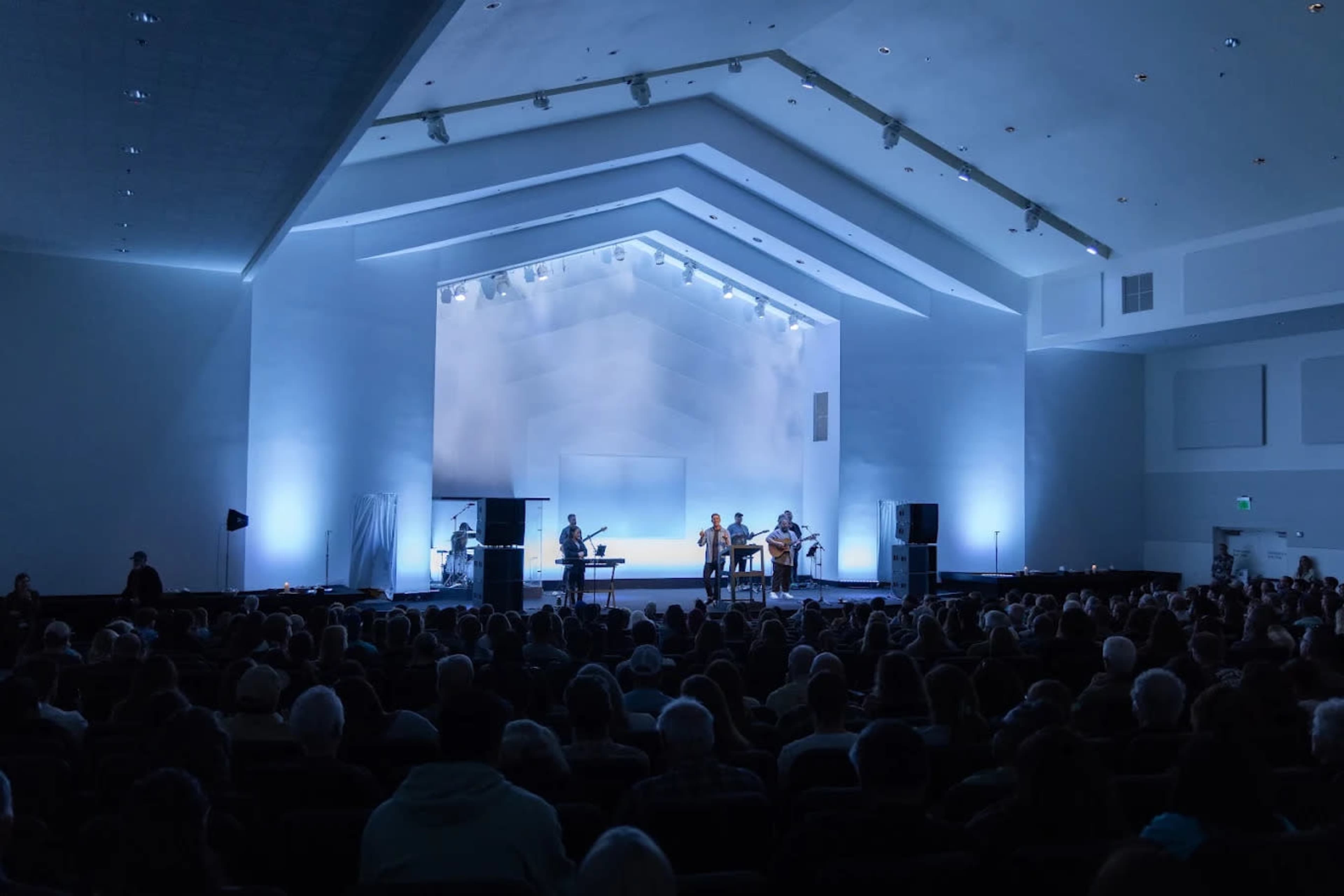 A band performs on stage in a well-lit auditorium filled with an audience.
