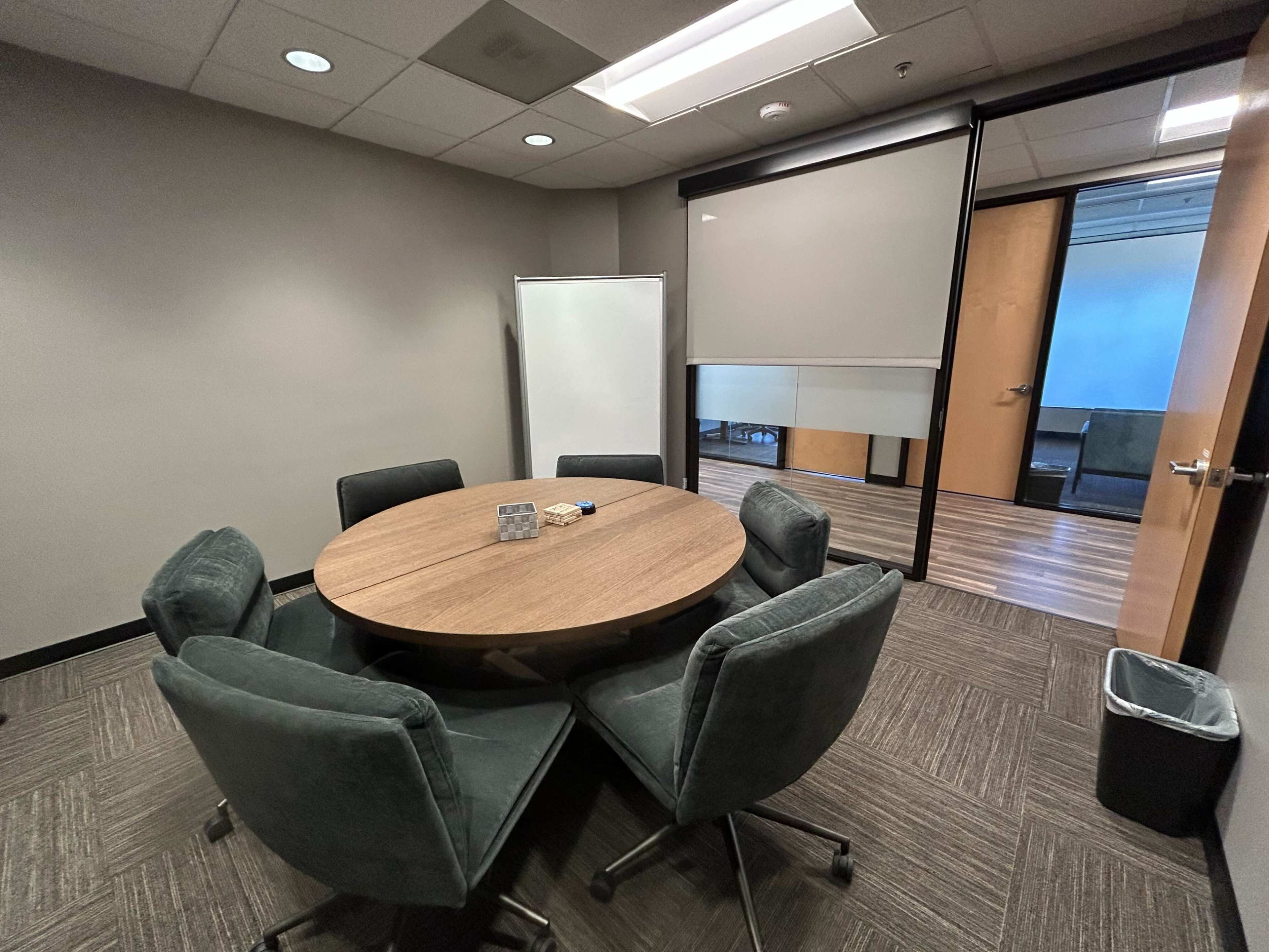 The image shows a small conference room with a round wooden table surrounded by six gray upholstered chairs, a whiteboard, and an office entrance in the background.