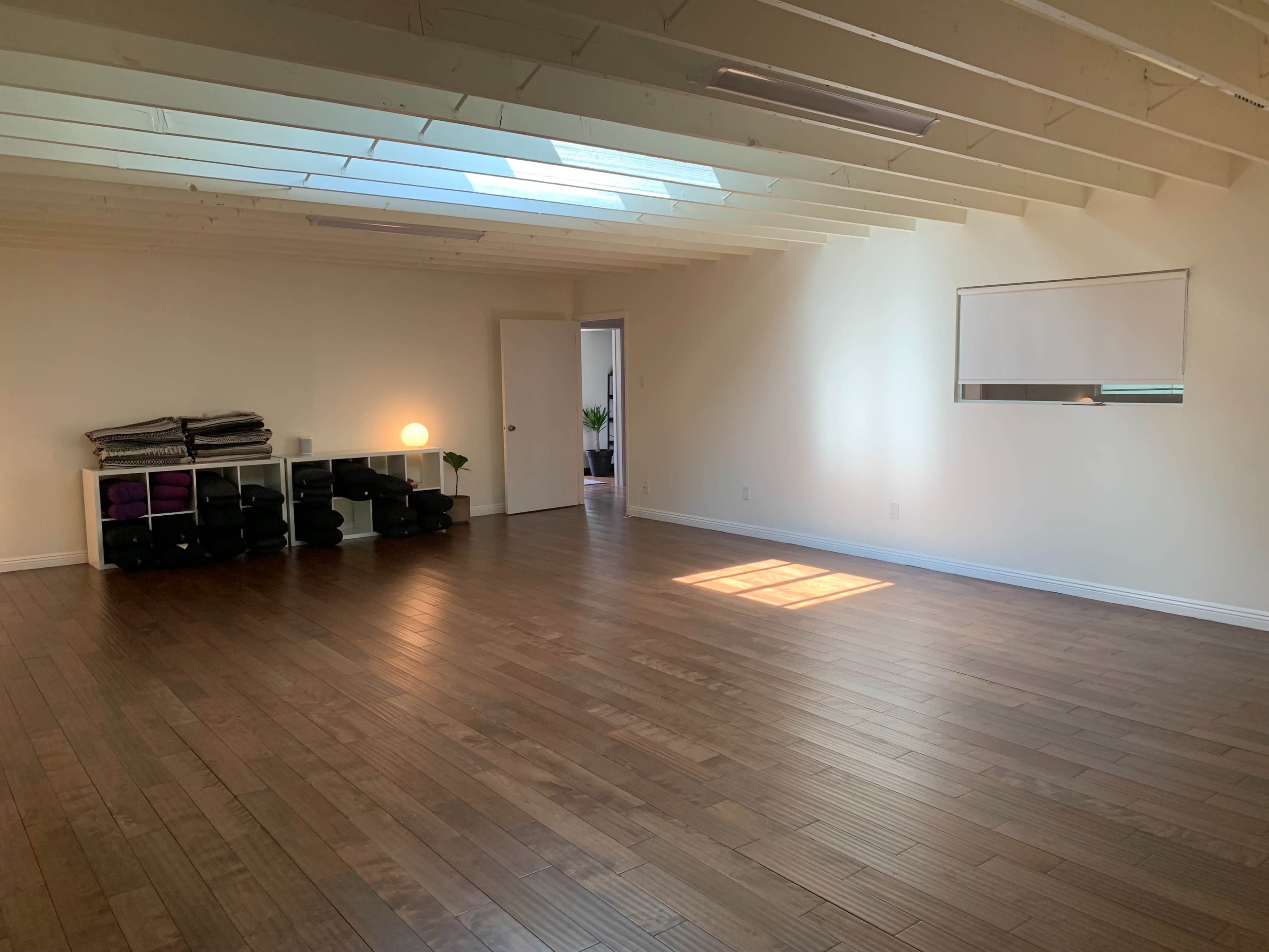 A spacious, empty room with wooden floors, a large window letting in light, and a shelf containing yoga mats and blocks.