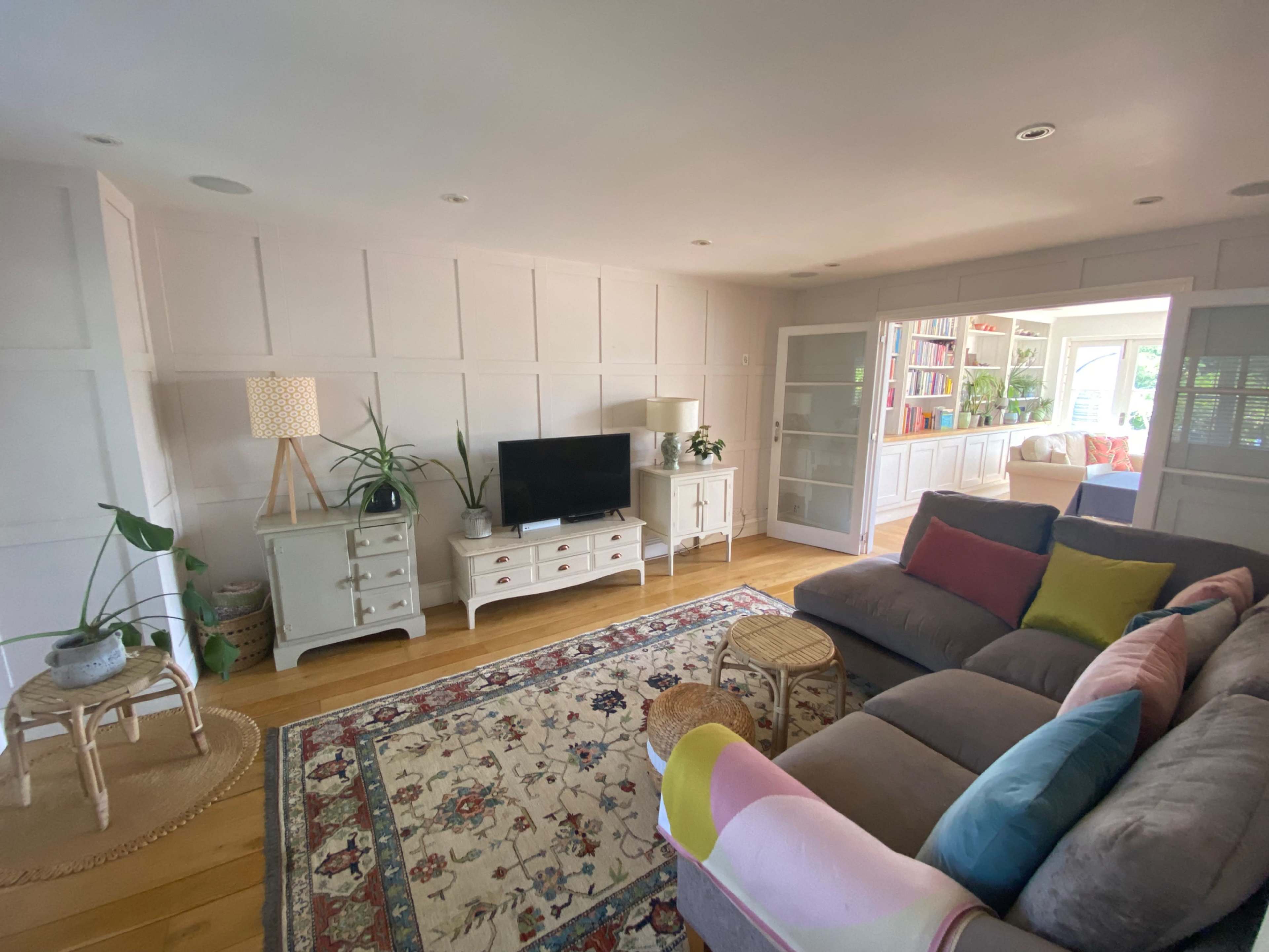 The image shows a bright living room featuring a sectional sofa, a coffee table on a patterned rug, and a television on a white media console, with decorative plants and bookshelves in the background.