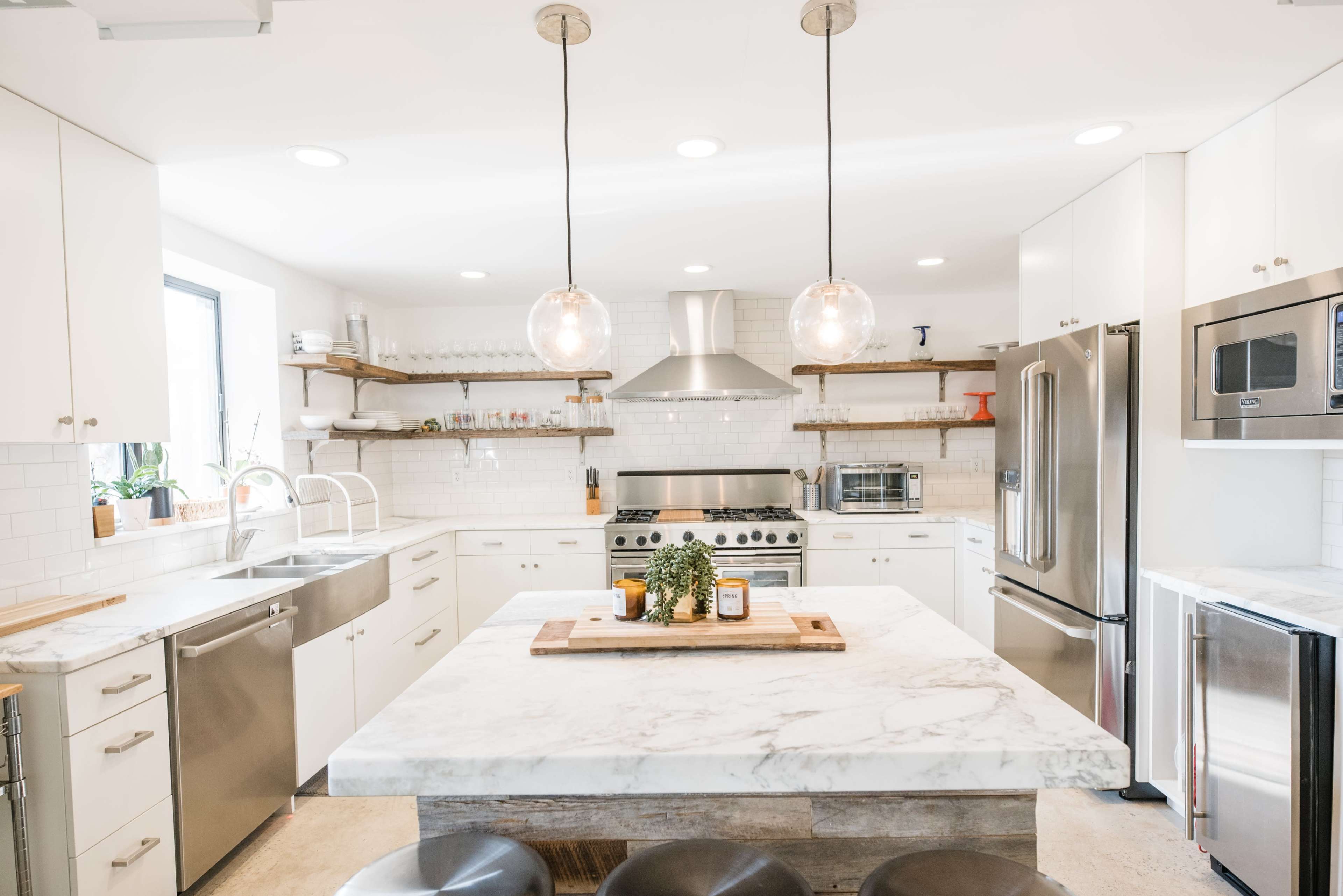 A modern kitchen features white cabinetry, a marble island, stainless steel appliances, and open shelving with light fixtures hanging above.