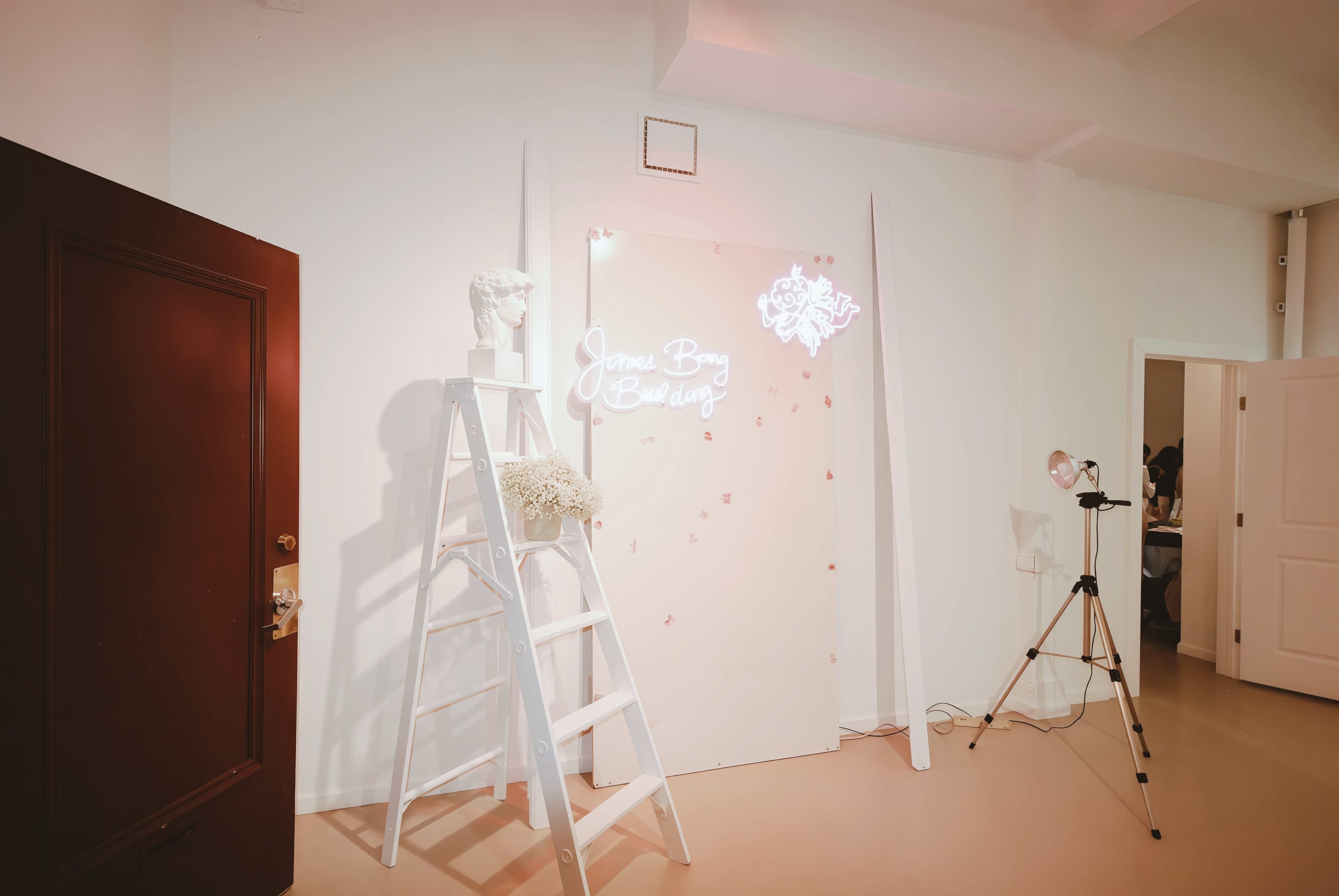 The scene features a minimalist interior with a pink backdrop adorned with decorations, a white ladder, a standing lamp, and a decorative bust statue.