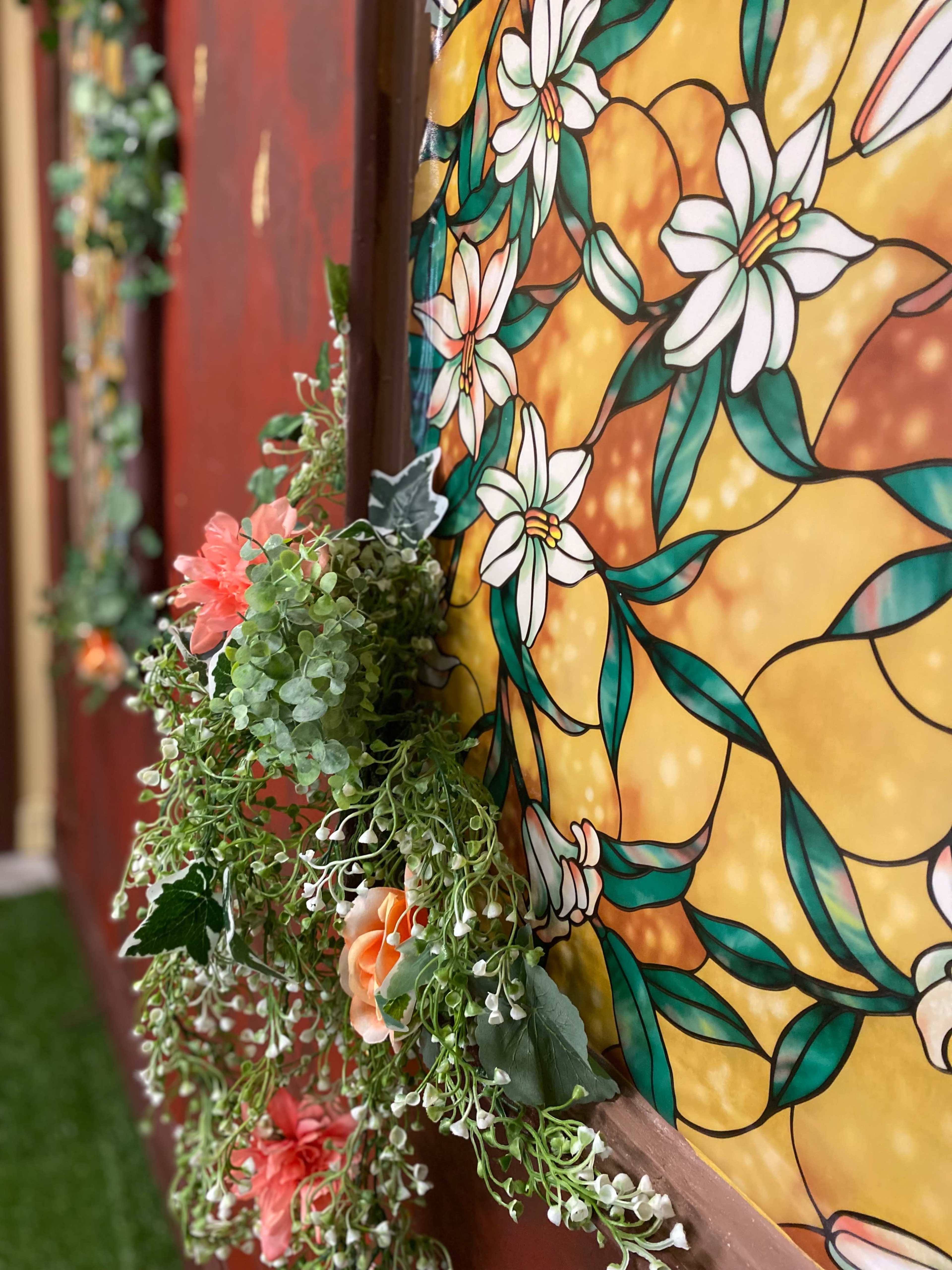 The image features a close-up of a decorative wall adorned with stained glass depicting floral patterns and a garland of artificial greenery and flowers draped along the edge.