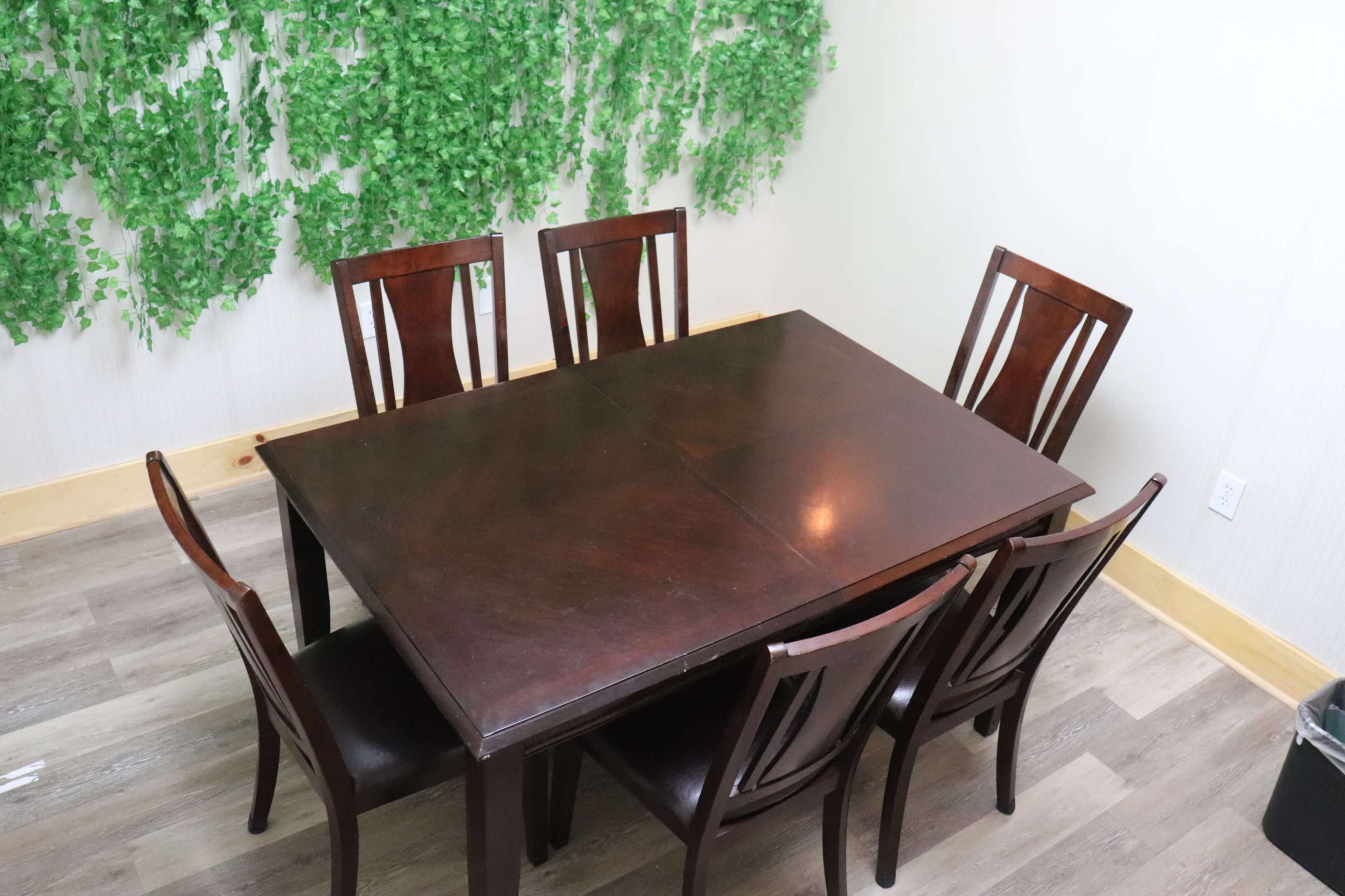 A dark wooden dining table with eight chairs sits in a room featuring a green vine backdrop against a light-colored wall.