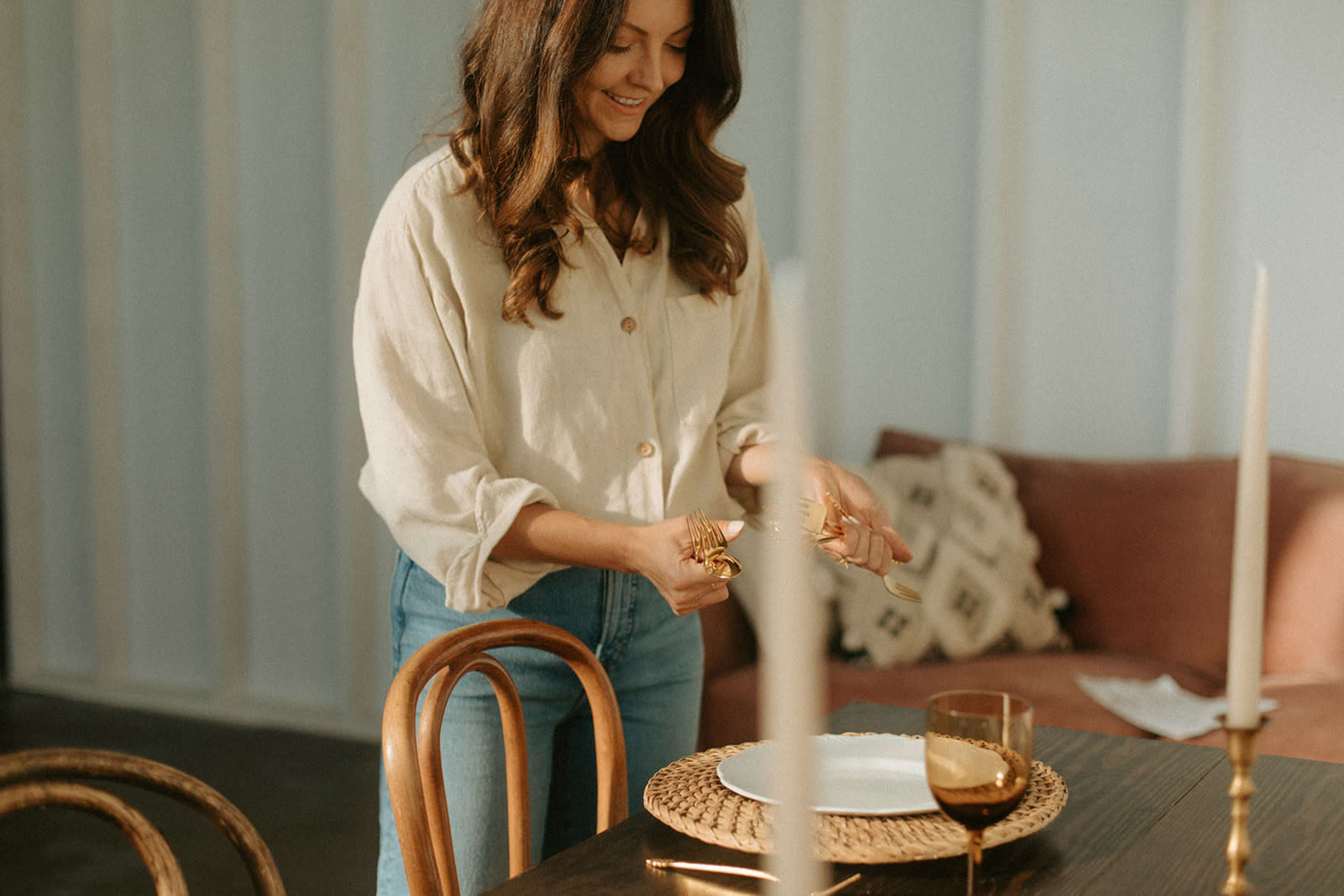 A woman sets a table for a meal, arranging utensils while standing next to a wooden dining table in a softly lit room.