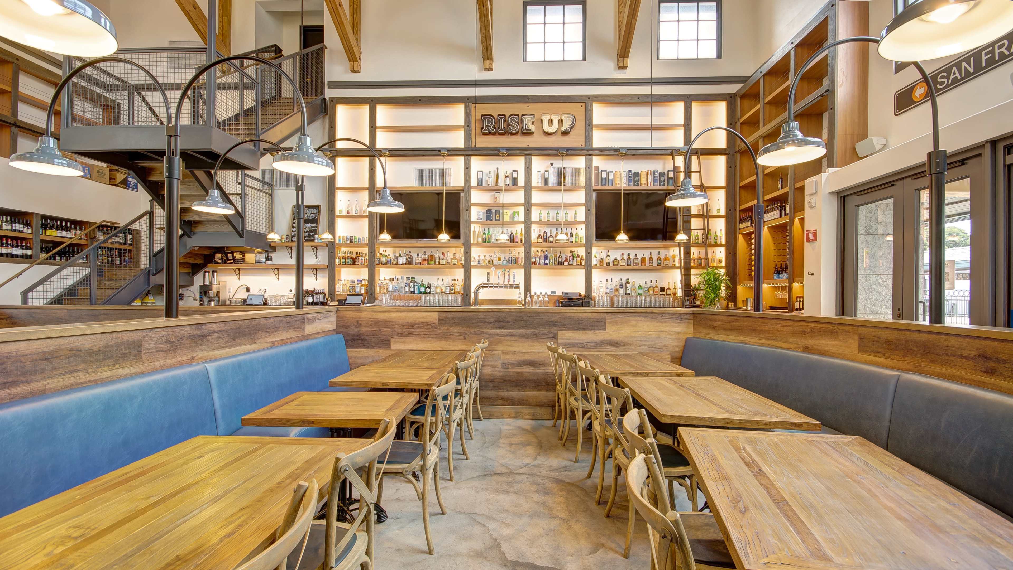 A contemporary restaurant interior with wooden tables, blue seating, and a well-stocked bar lined with bottles behind a wooden counter.