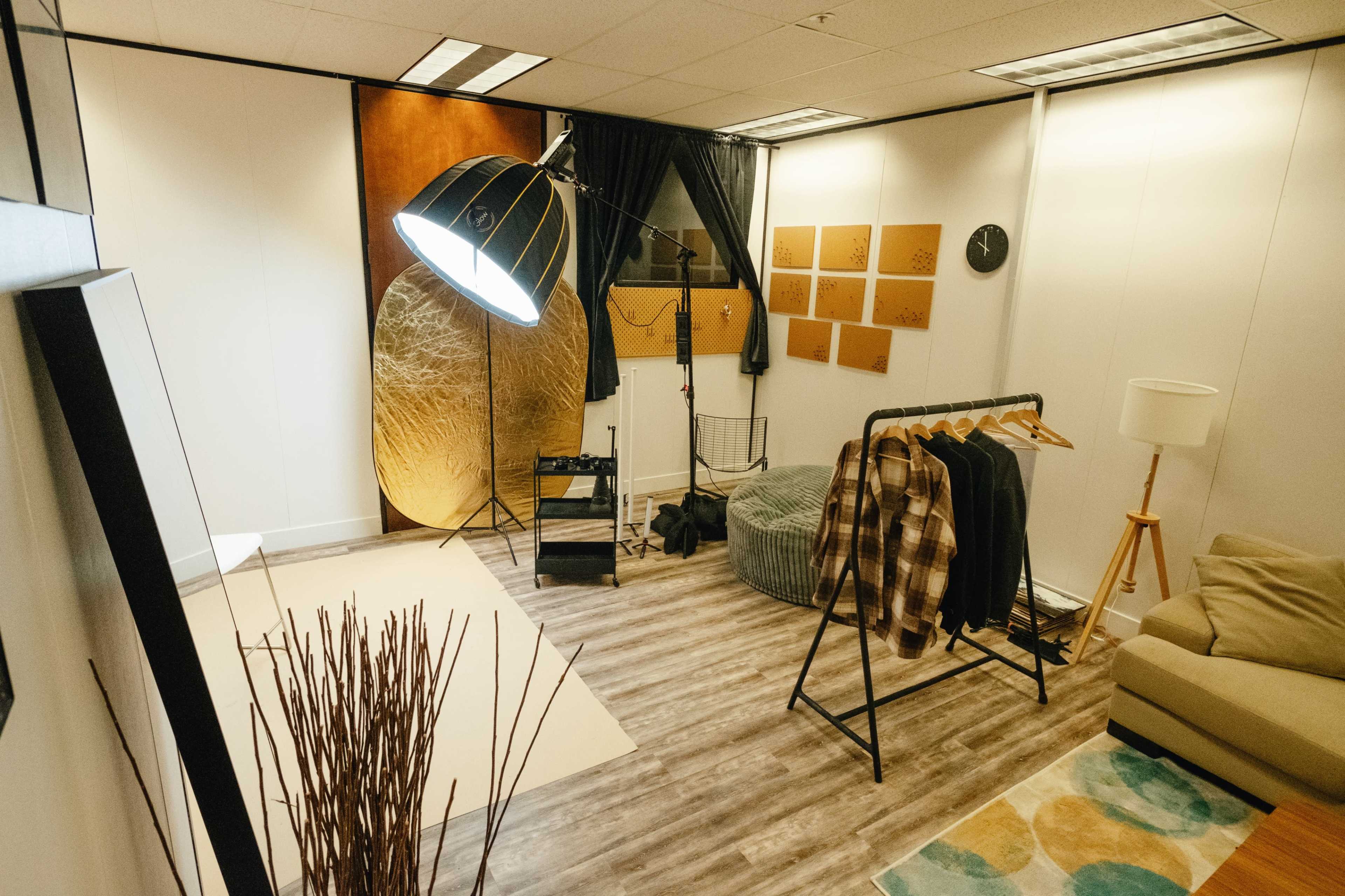 A modern studio space features a seating area, clothing rack, a large floor lamp, and a backdrop with textured panels.