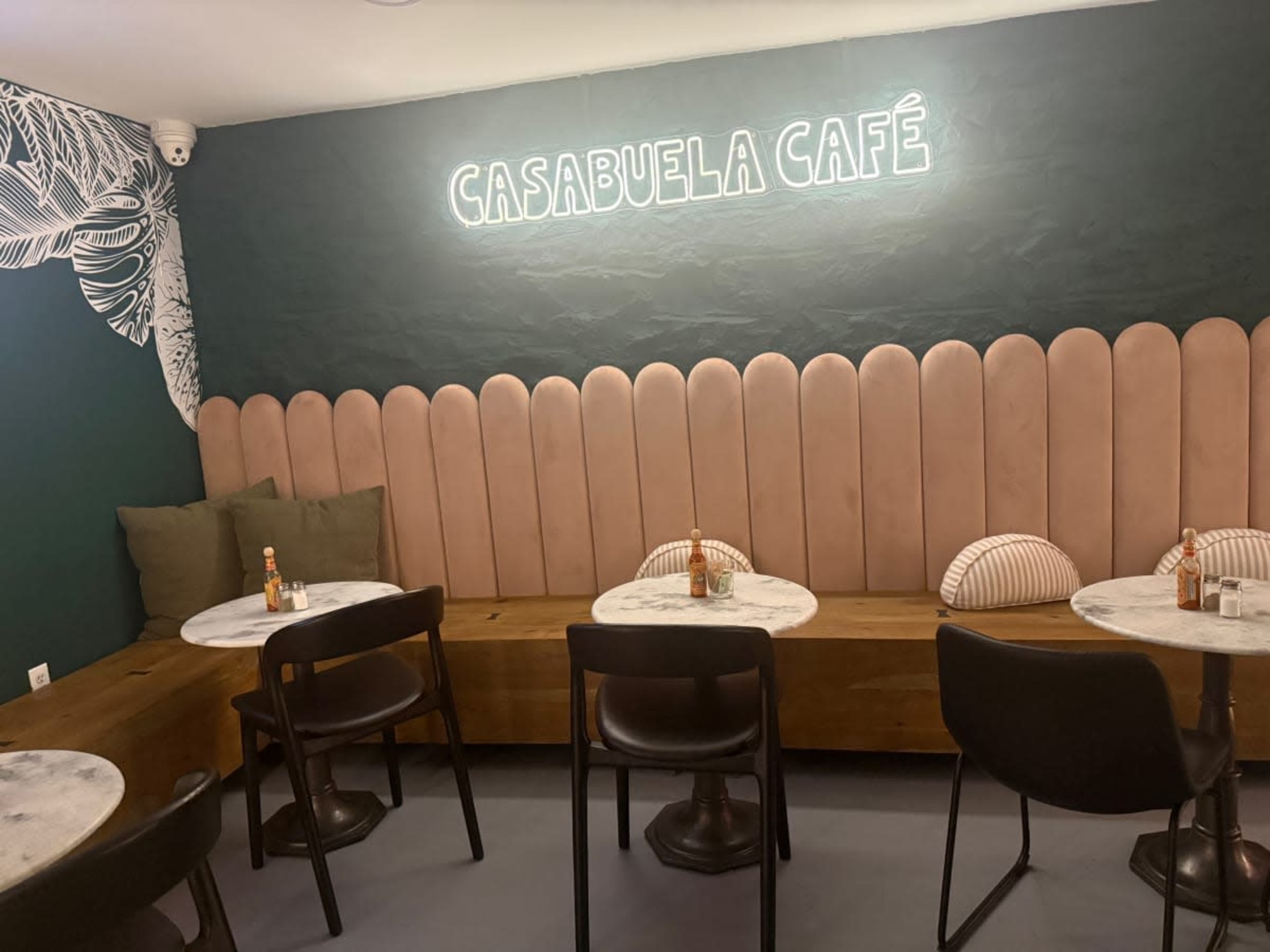 The image shows a cozy café interior with a neon sign that reads "CASABUELA CAFÉ," featuring pink padded seating and small round tables.