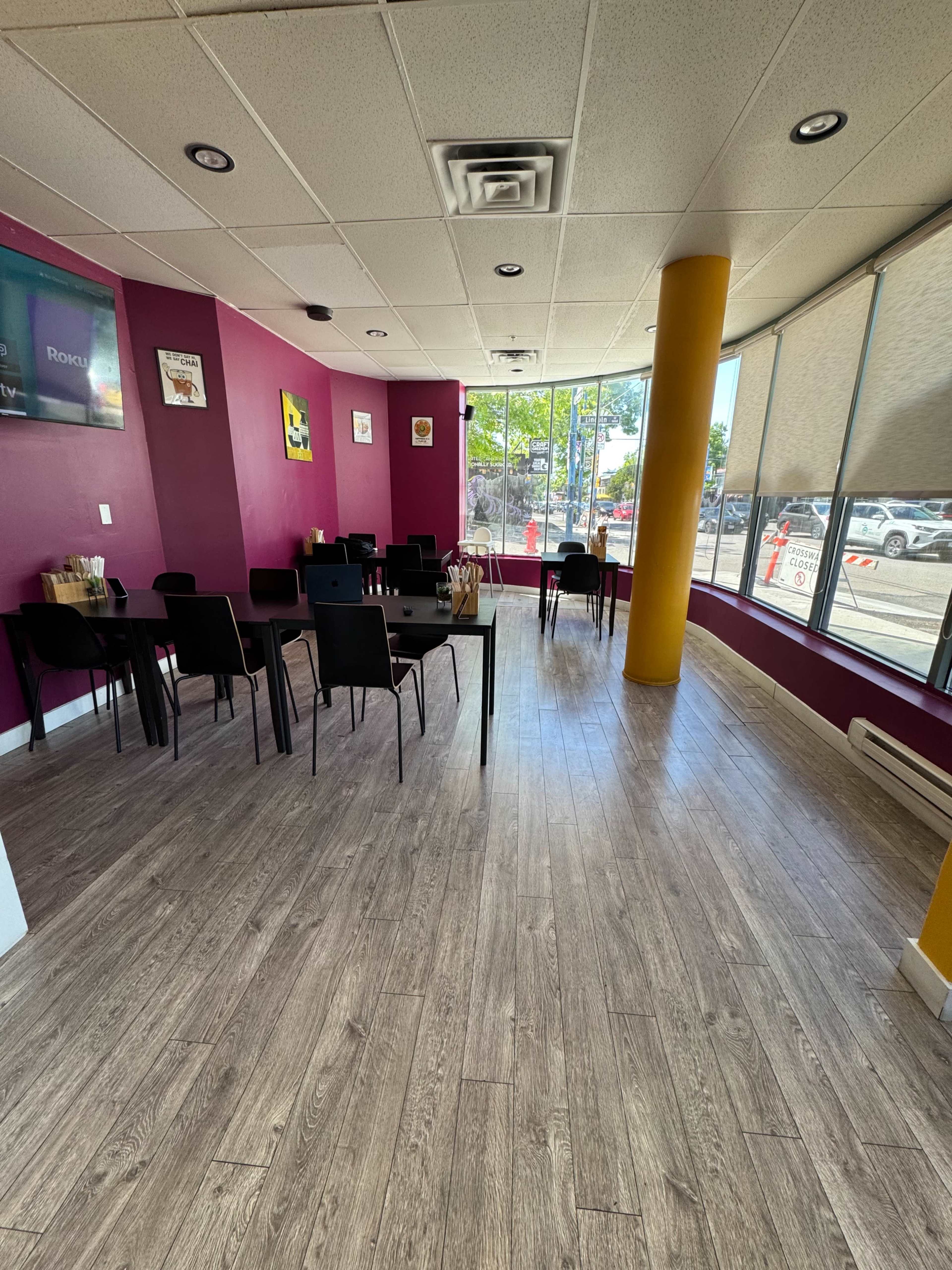 The image shows a brightly lit restaurant interior with purple walls, wooden flooring, and several black chairs and tables arranged neatly.