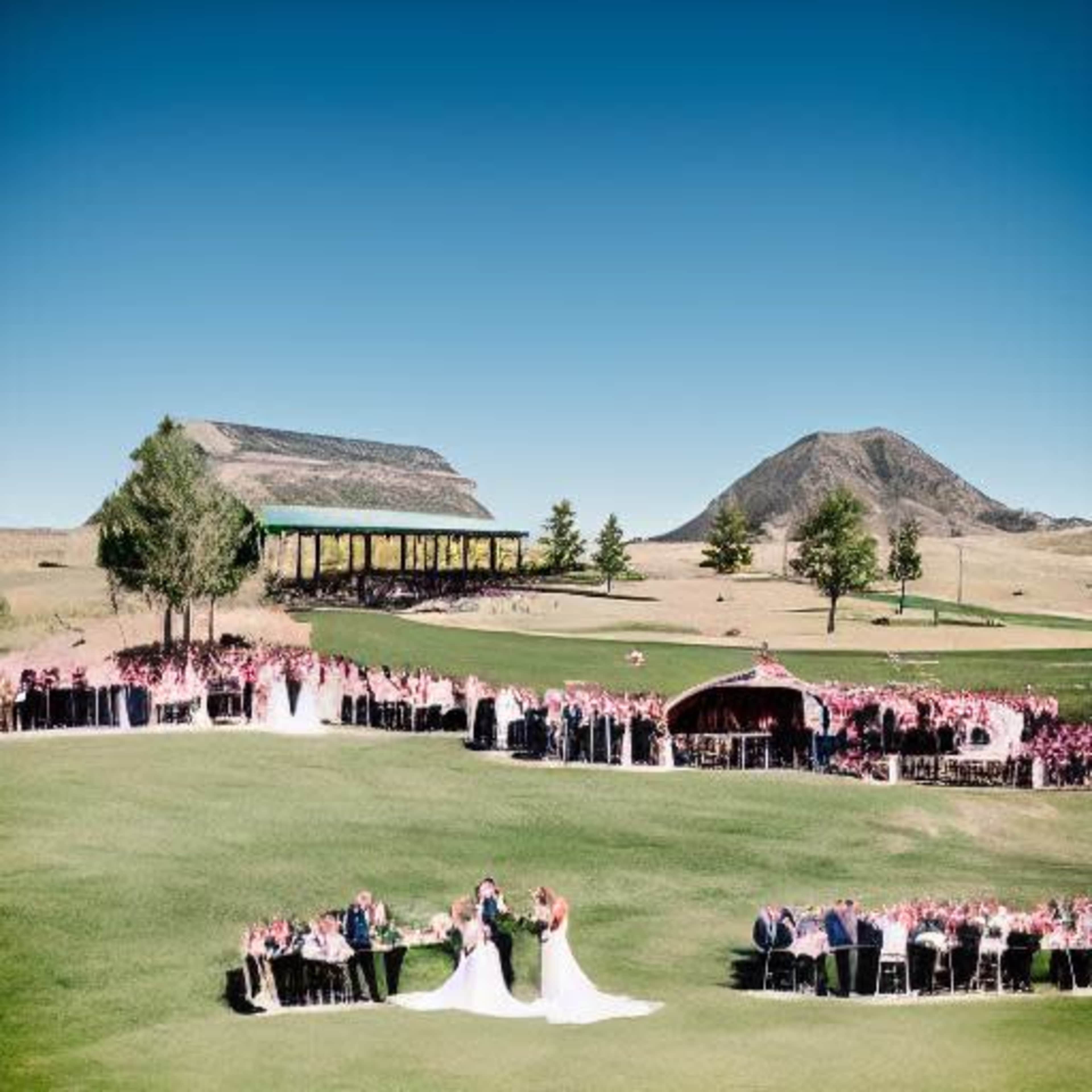 A large outdoor wedding ceremony is set on a grassy field with guests seated in rows and mountains in the background.