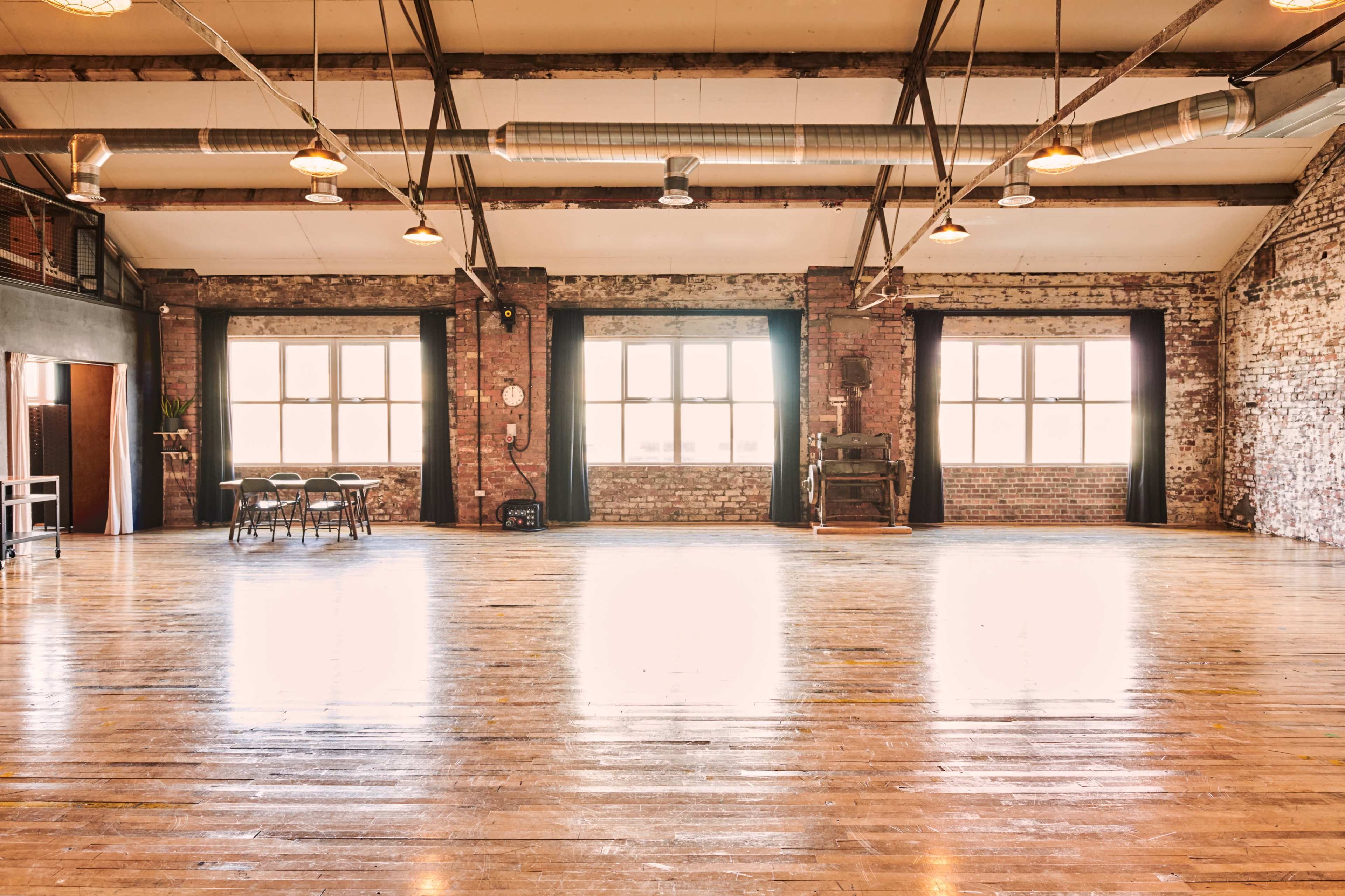 A spacious, industrial-style room with wooden floors, large windows, and exposed brick walls, featuring a few chairs and a table arranged in the center.