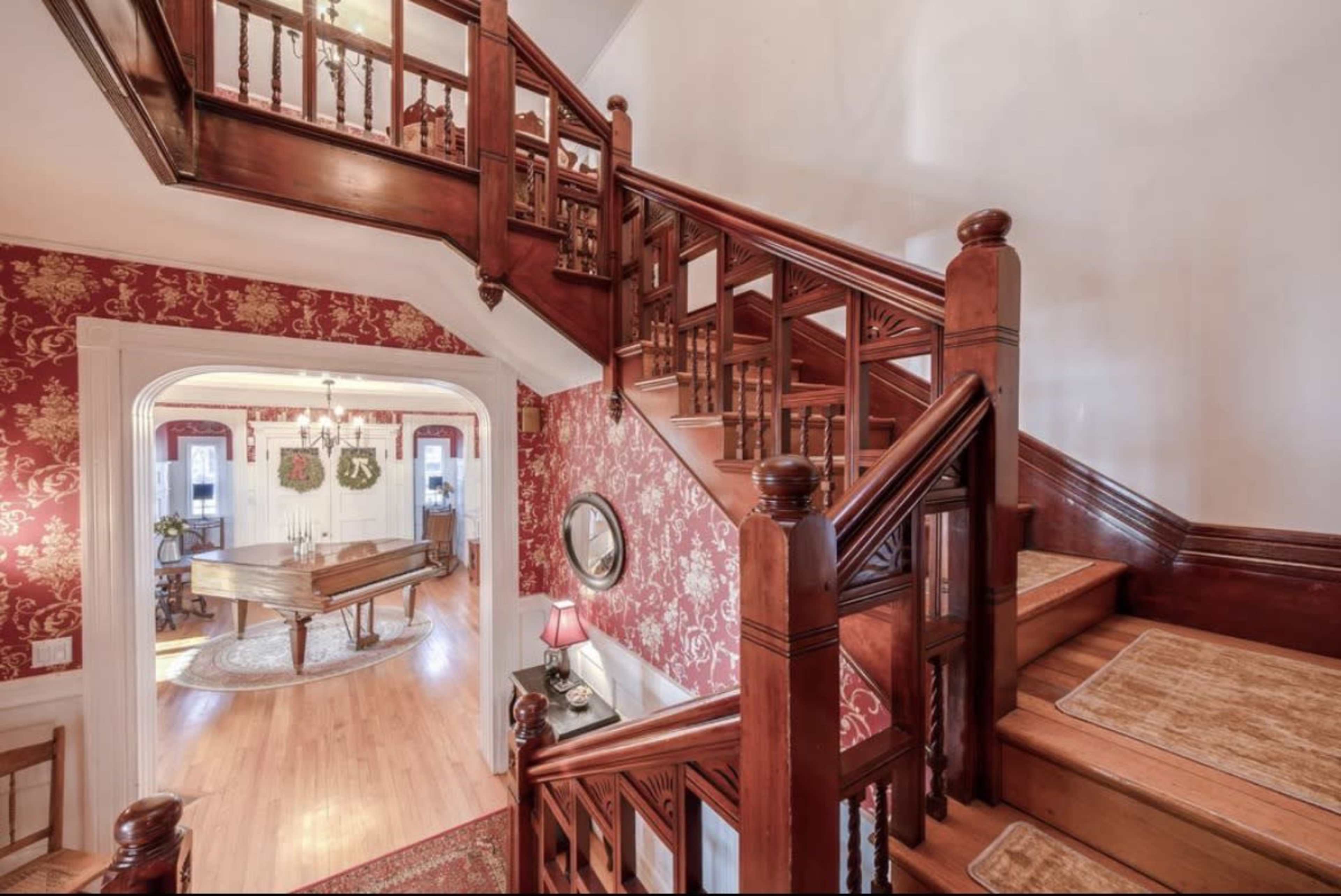 A wooden staircase with intricate railings leads to an upper level, while opulent wallpaper adorns the foyer and a grand piano is visible in the background.