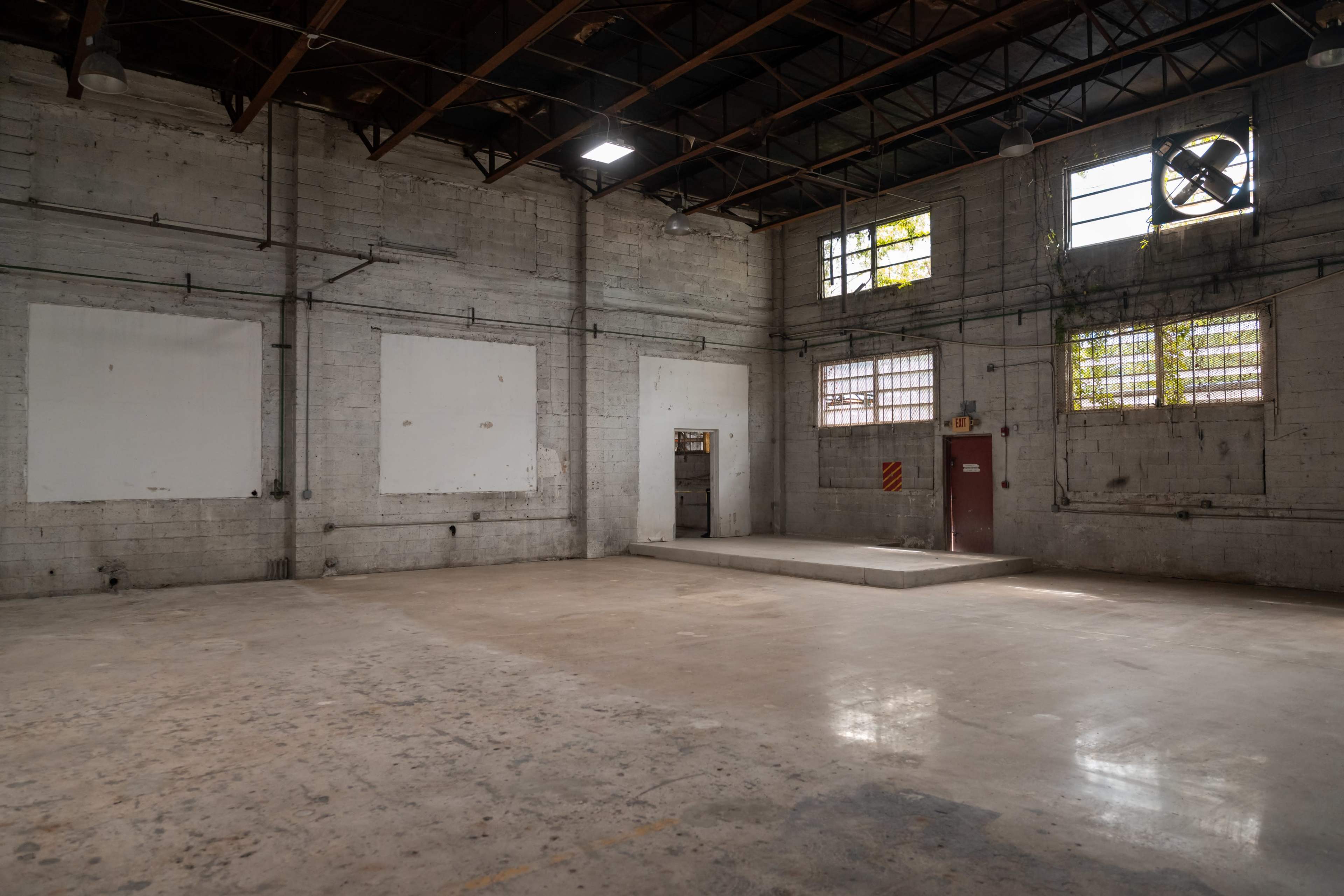The image shows an empty industrial warehouse with concrete walls, large windows, and a raised platform at one end.