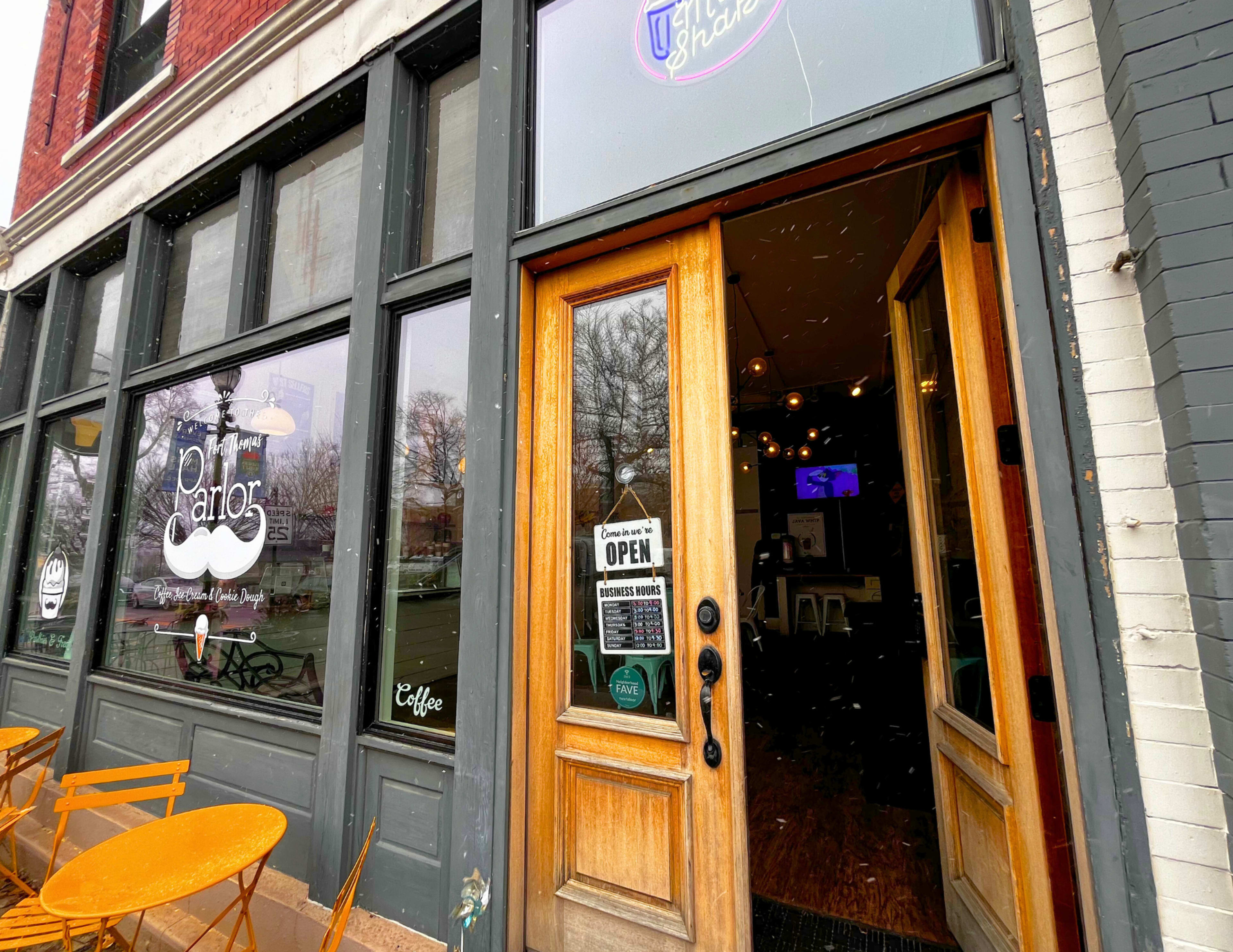 The image shows the entrance of a café with wooden doors, large windows, and outdoor seating.