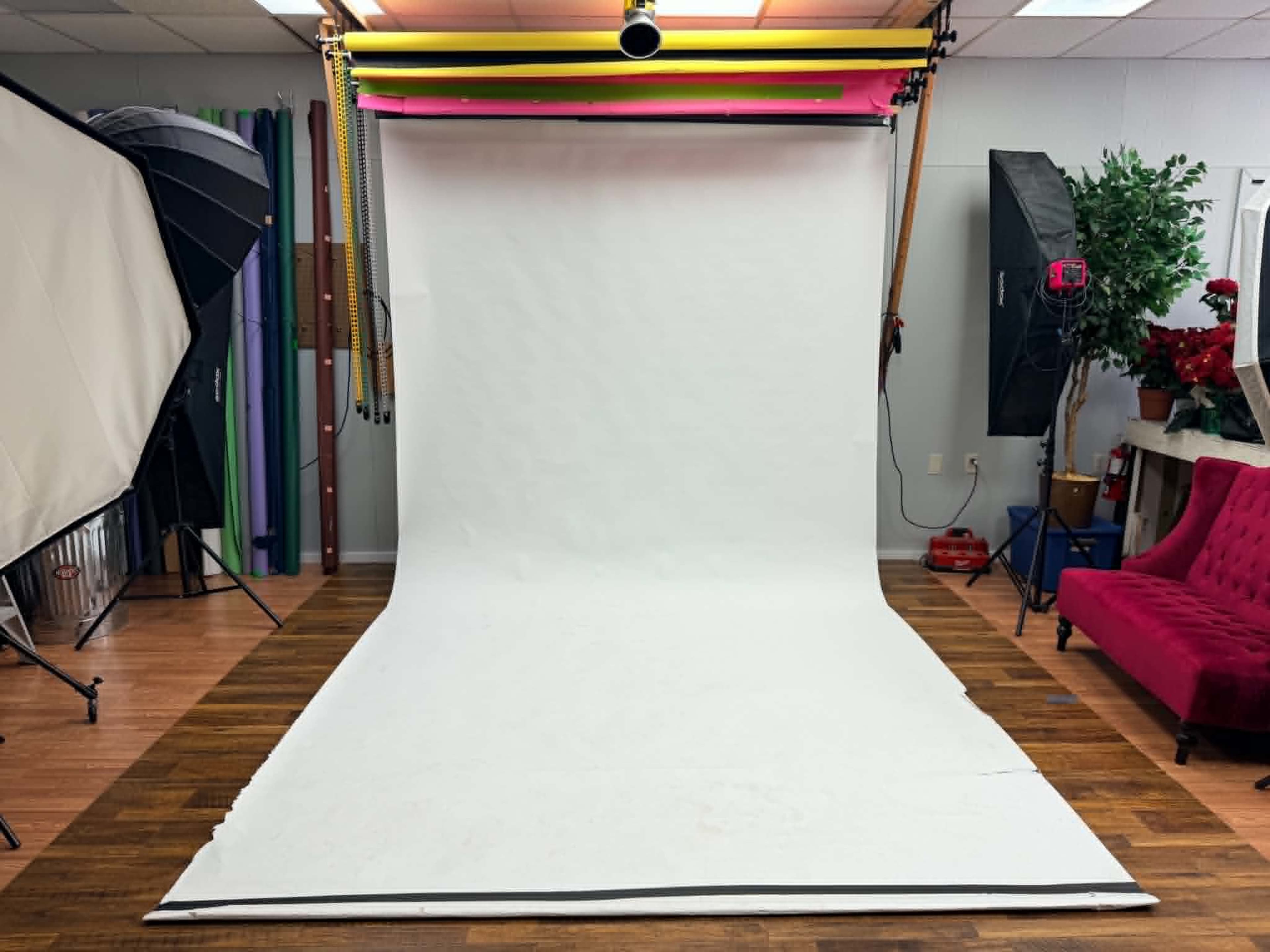 The image shows a photography studio with a large white backdrop, studio lighting equipment on both sides, and colorful rolls of background paper hanging from a rack.