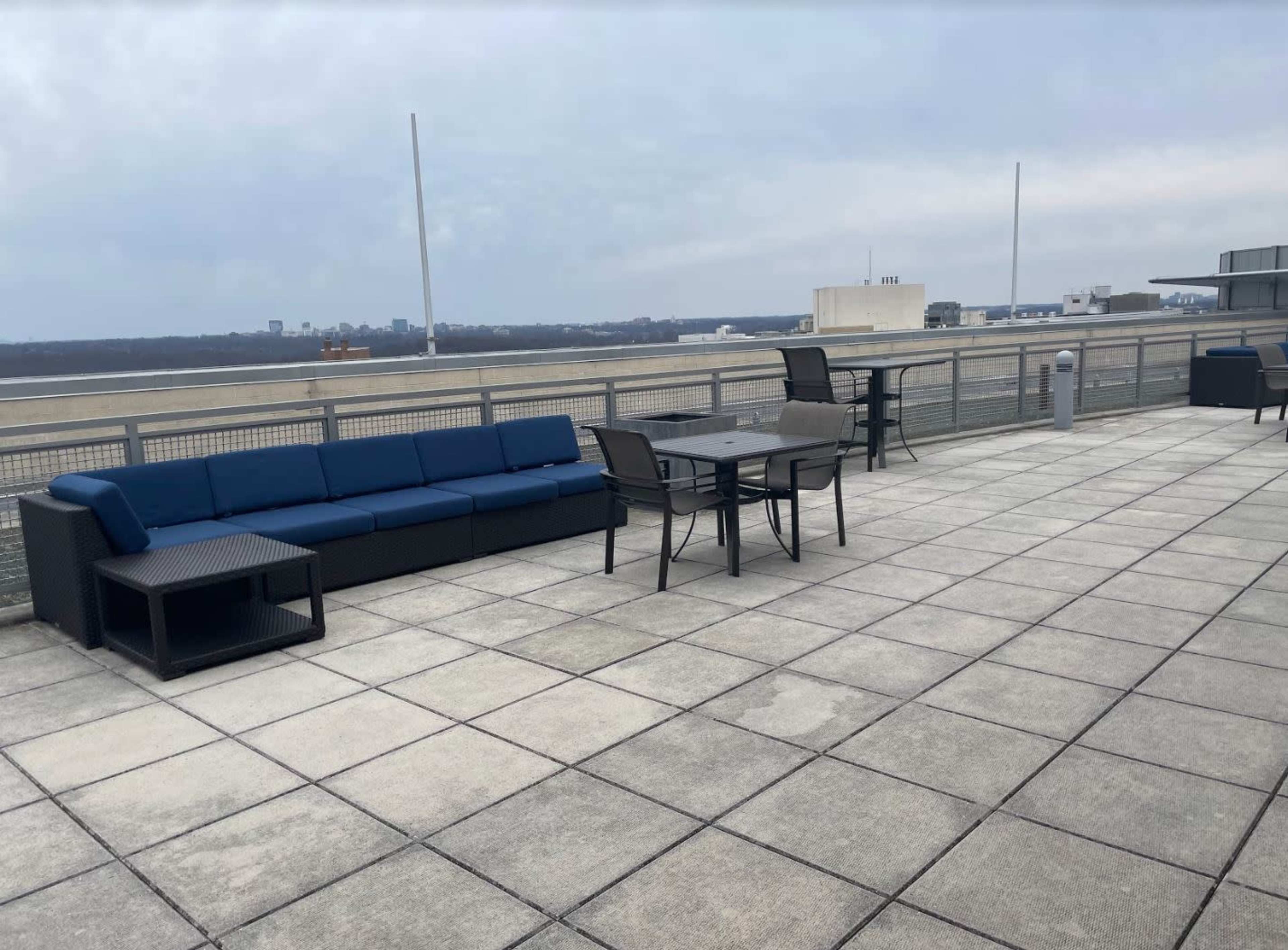 A rooftop terrace featuring a blue sectional sofa, a dining table with chairs, and a view of a distant city skyline.