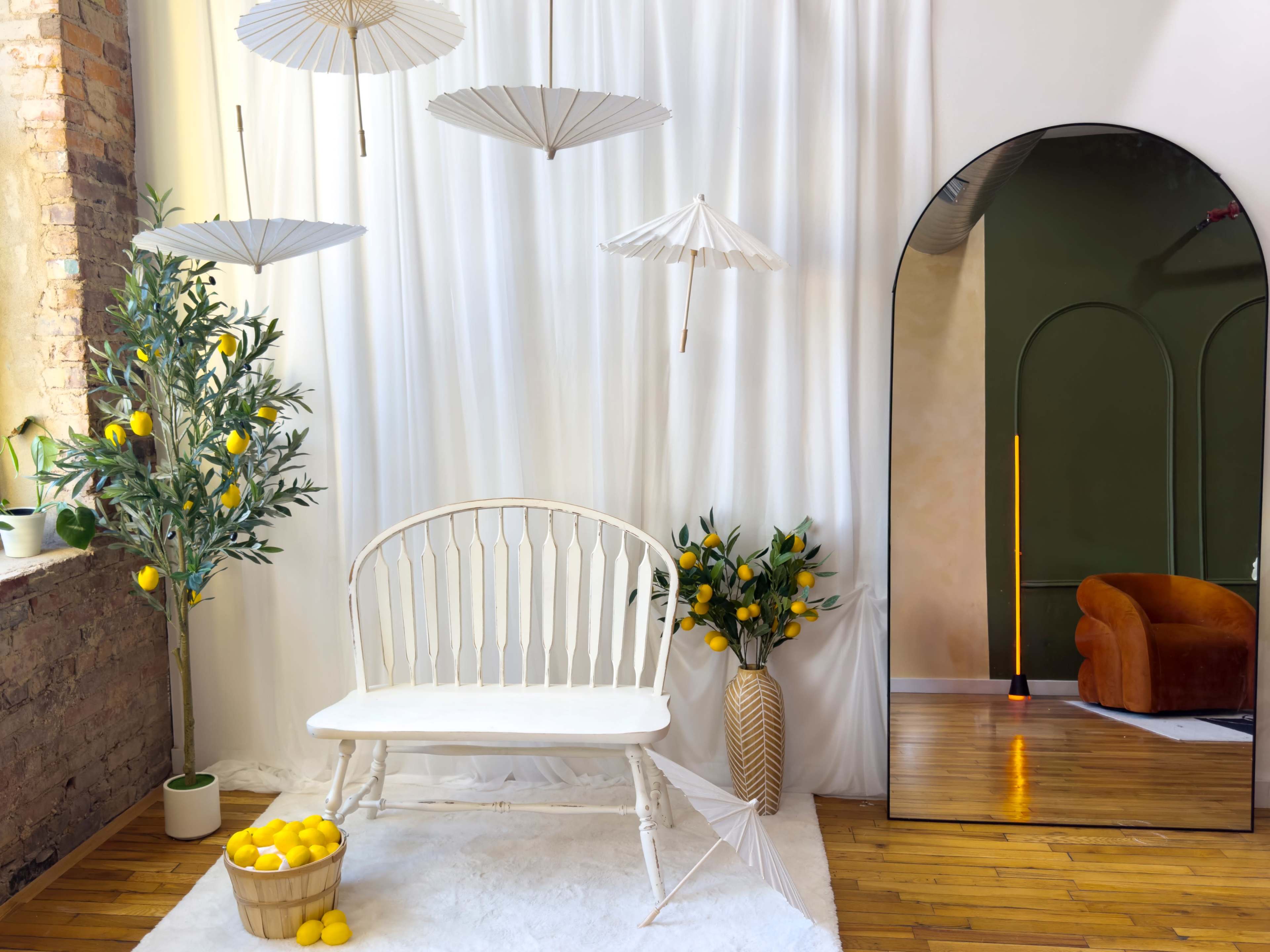 A white bench sits under hanging umbrellas, surrounded by decorative lemon trees and a large mirror reflecting a green wall and a brown couch.